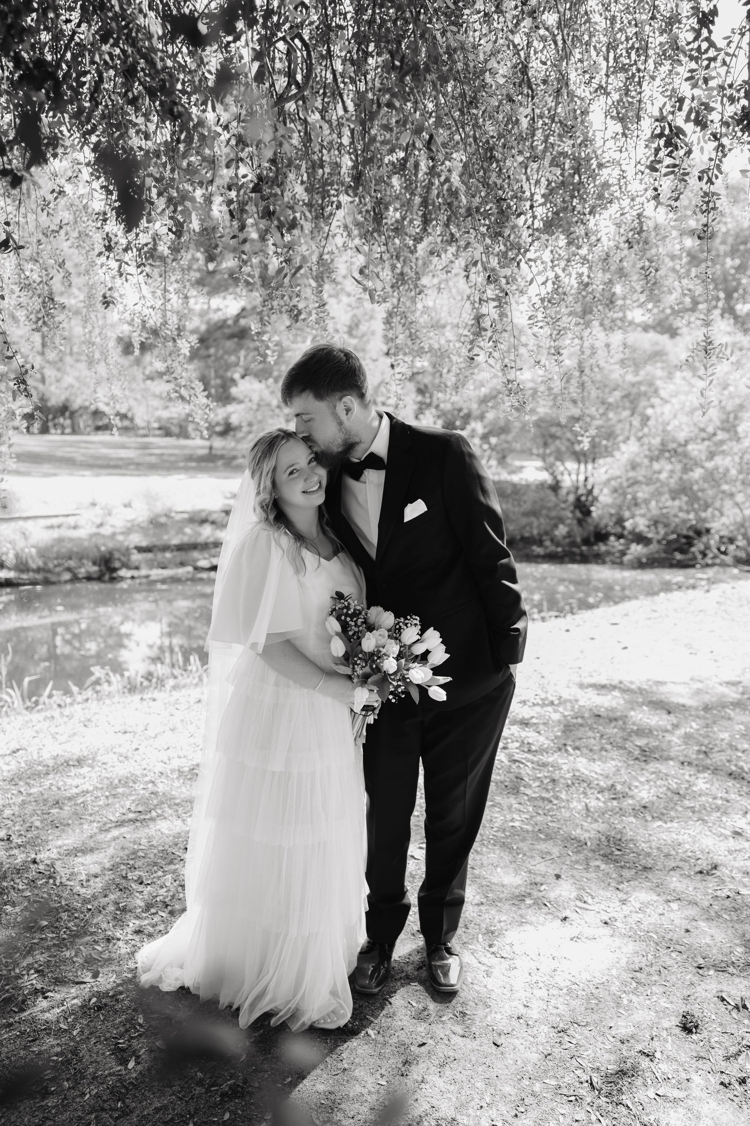 Black and white photo of bride and groom embracing under weeping willow tree by pond.