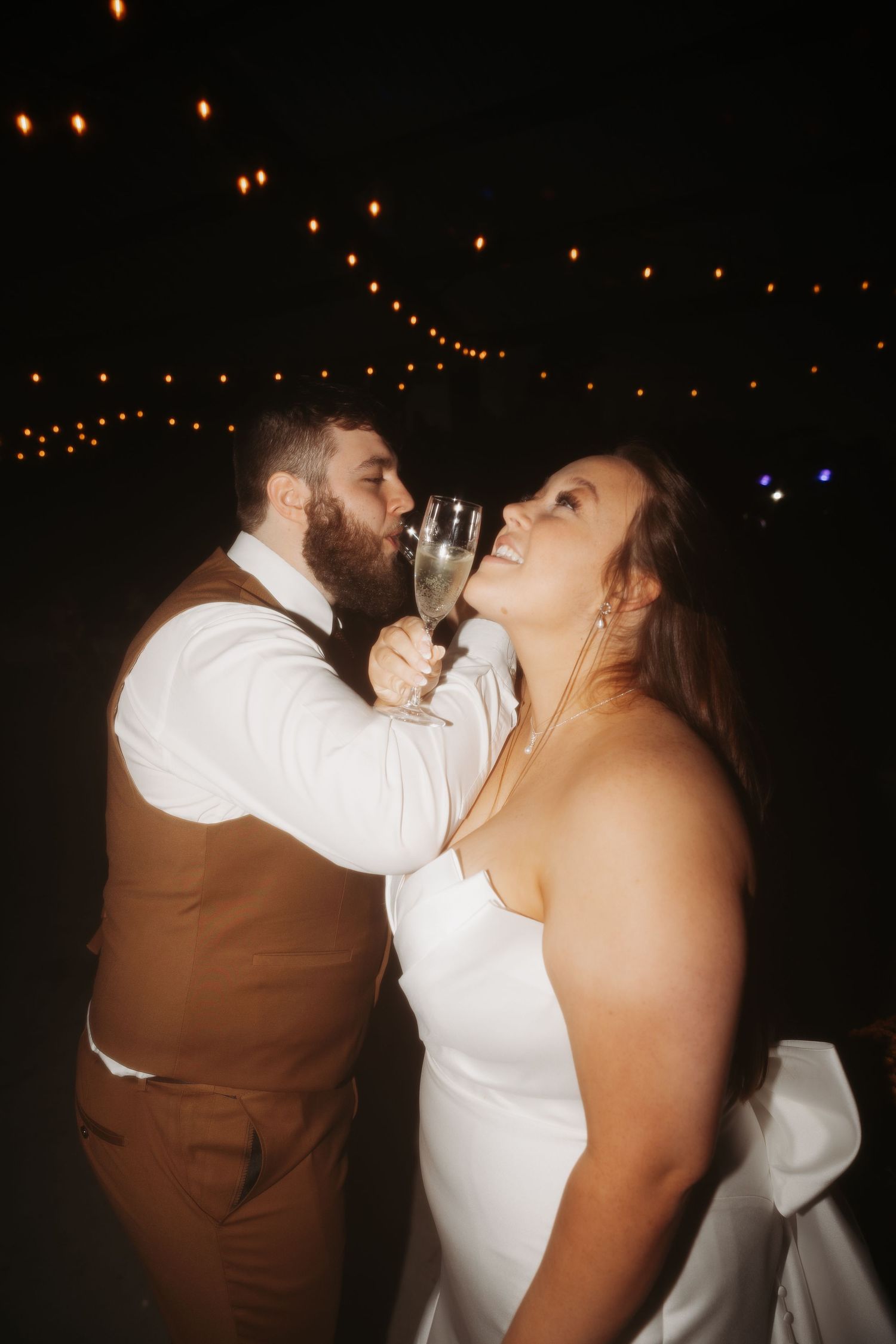 Bride and groom share a champagne toast under string lights during their evening wedding reception.