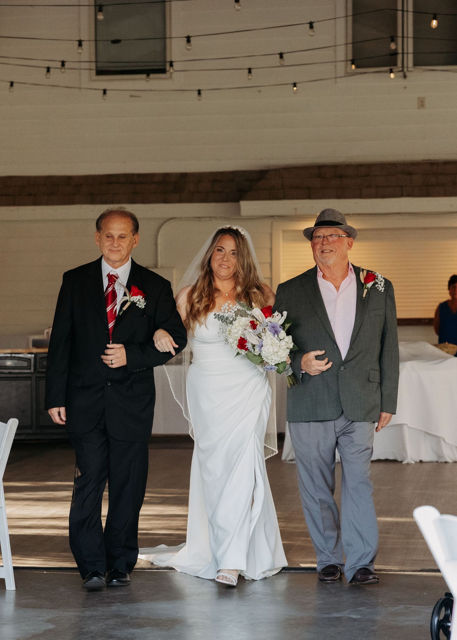 Bride walks down aisle escorted by two men in suits at indoor wedding venue with string lights.