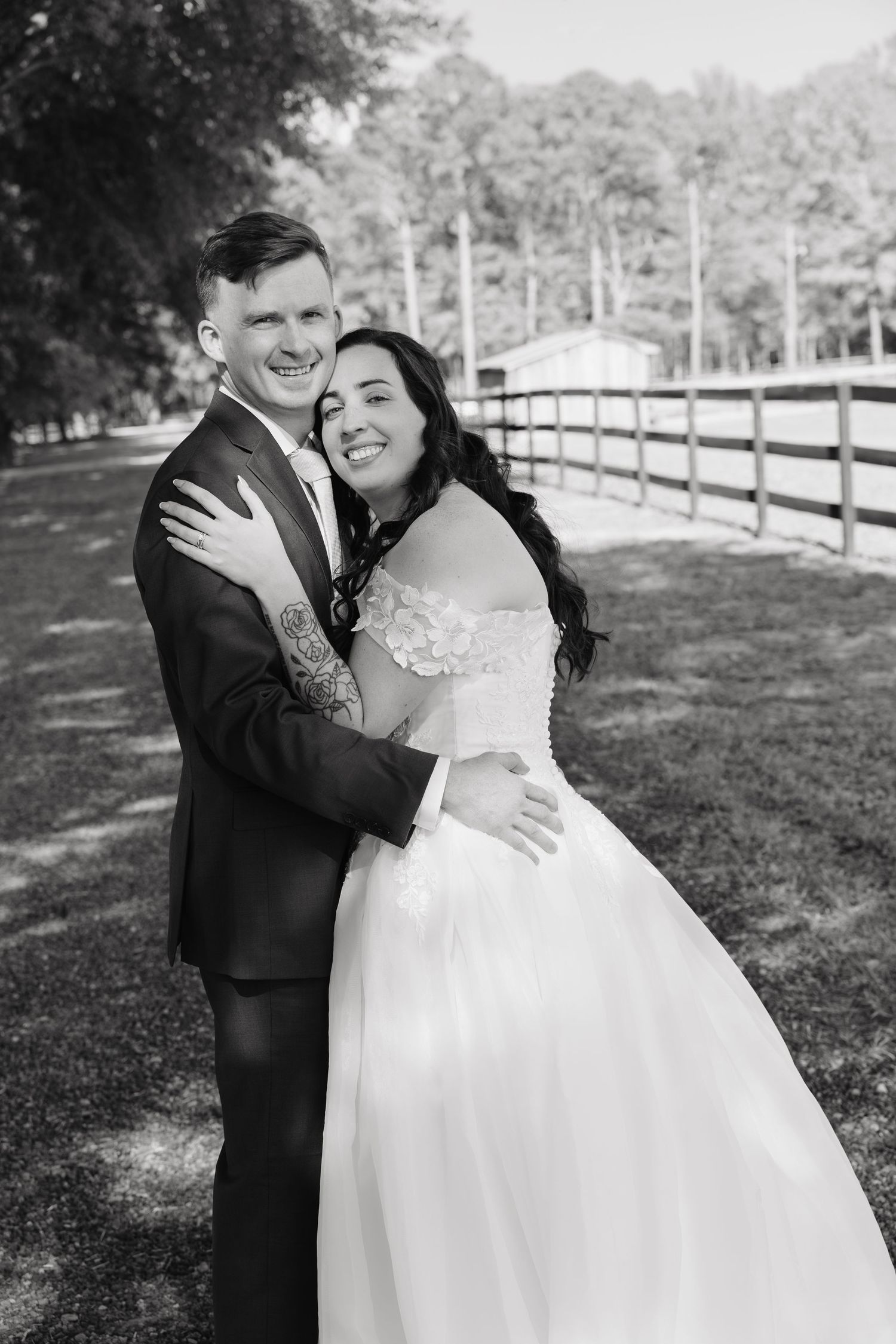 Happy bride and groom embrace outdoors near wooden fence in black and white wedding portrait.