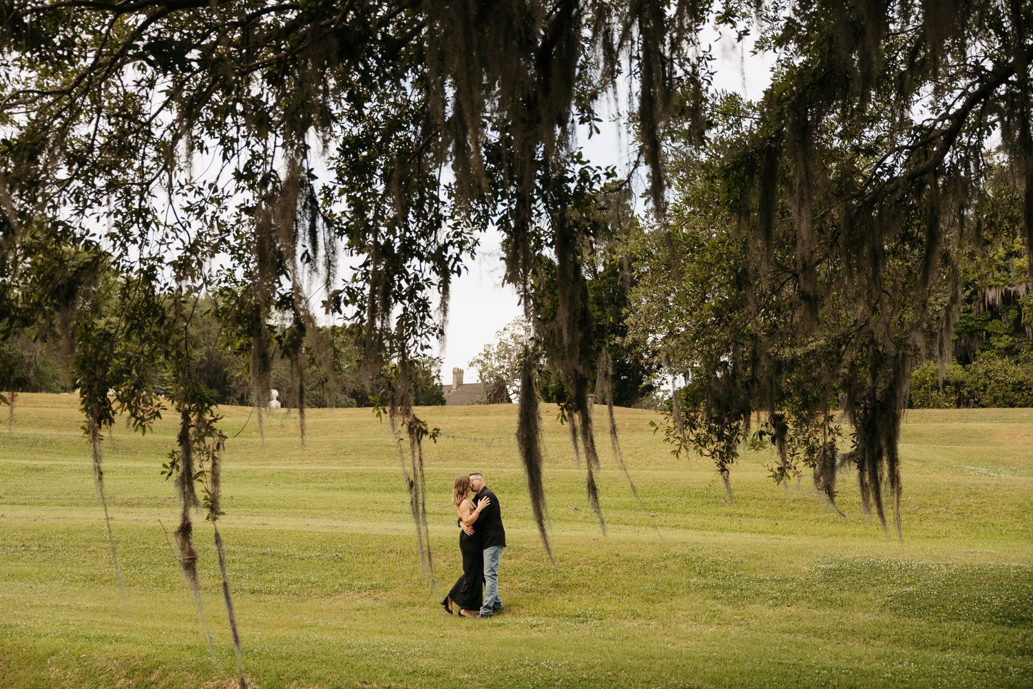Couple embracing on a lush green lawn beneath Spanish moss-draped trees during a romantic outdoor engagement session.