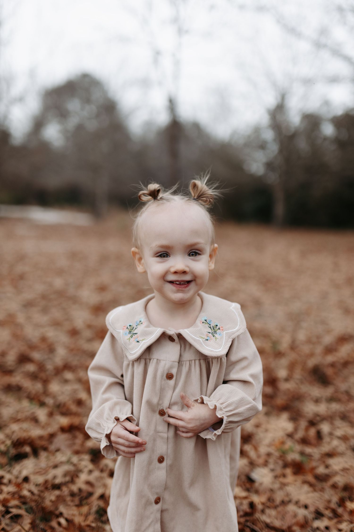 A child in a beige coat stands in a field of fallen autumn leaves with a joyful expression.