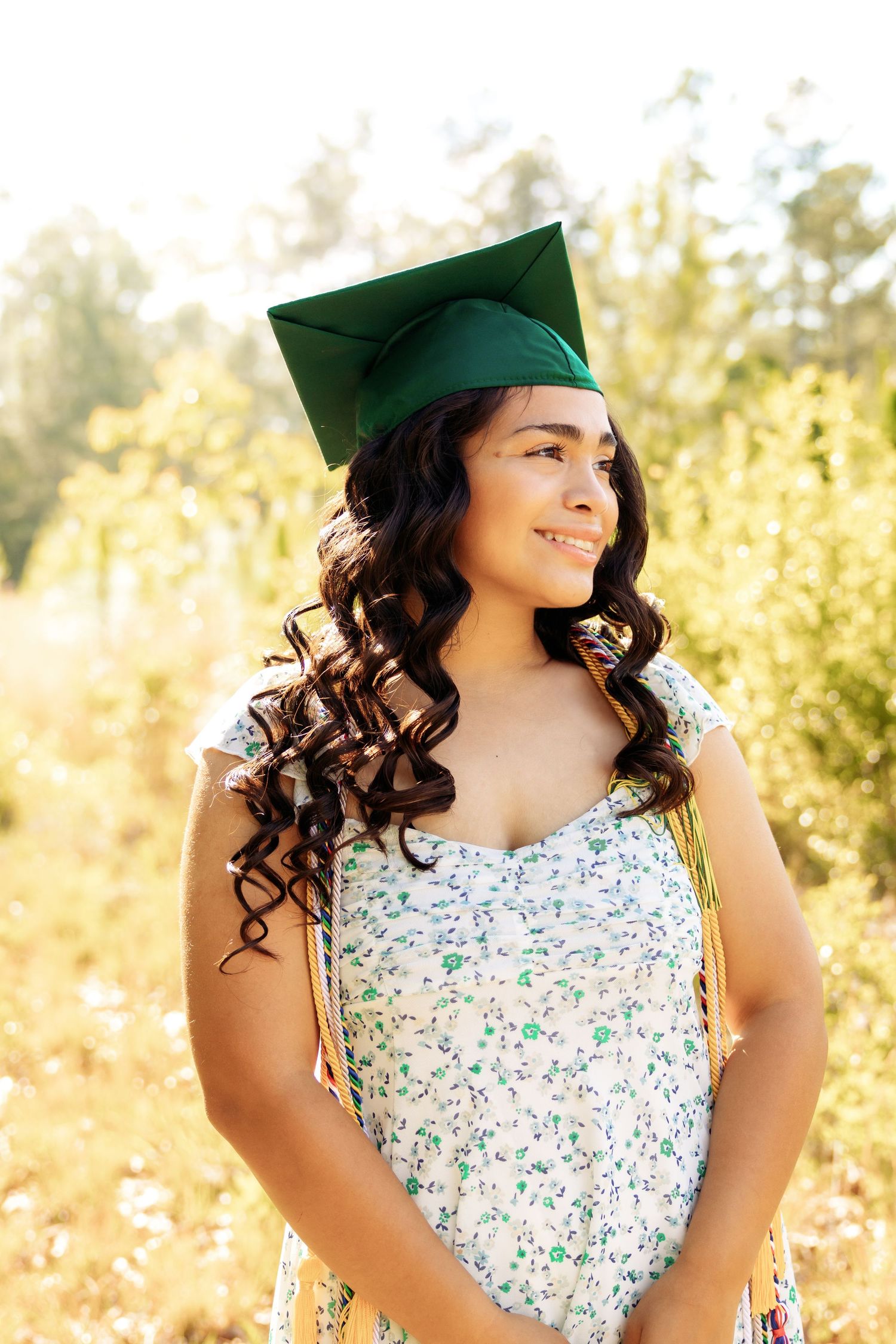 Graduate in green cap and floral dress smiles warmly in sun-drenched outdoor portrait.