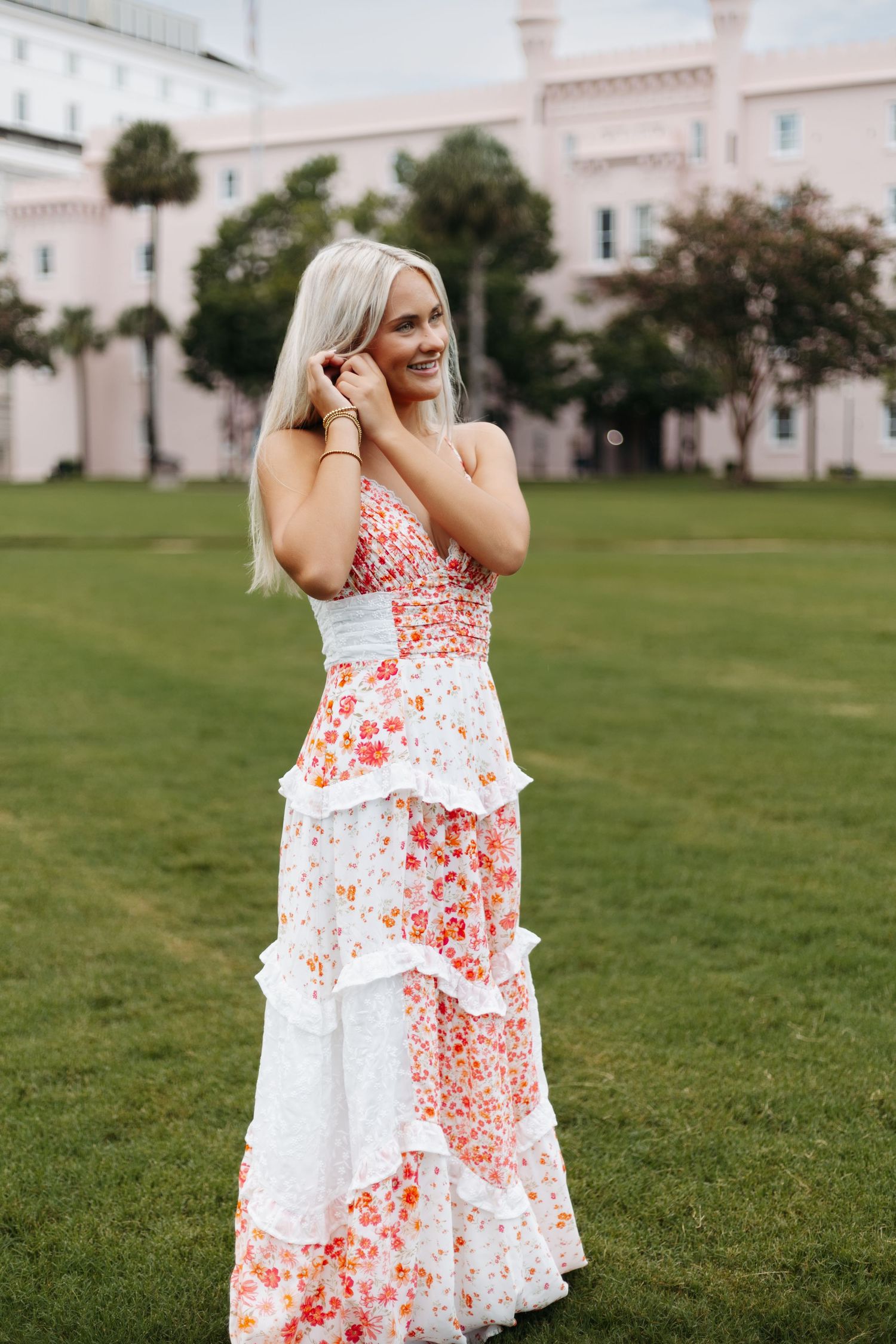 A floral tiered maxi dress in coral and white is worn outdoors on a grassy lawn with pink buildings in the background.