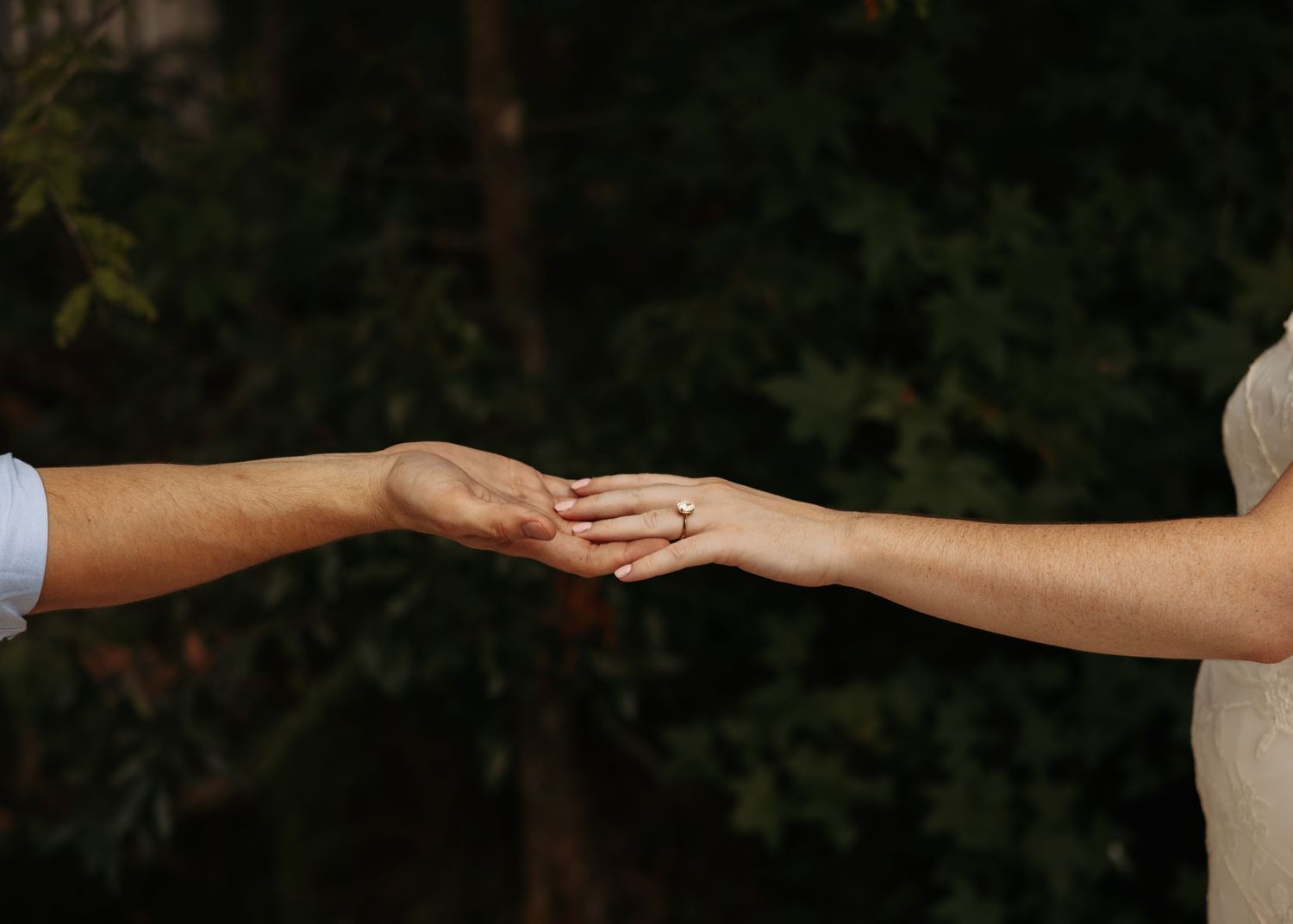 Two people gently holding hands outdoors, with a diamond engagement ring visible on the woman's finger.