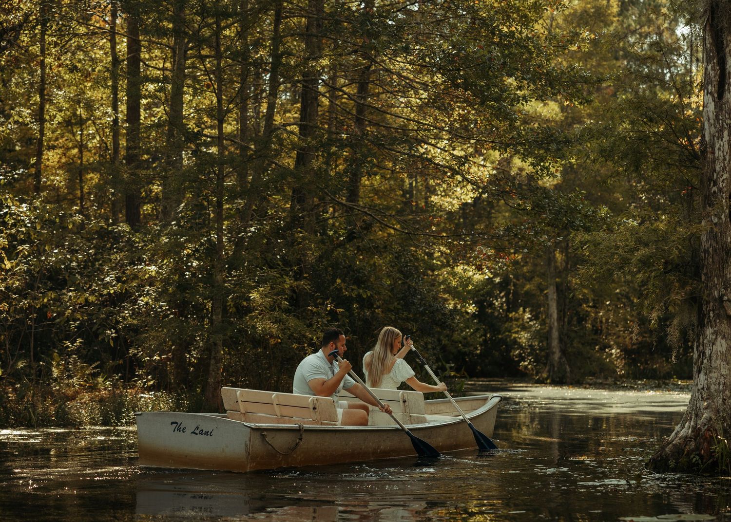 Couple rowing a wooden canoe on a serene forest river surrounded by lush green trees in soft natural light.