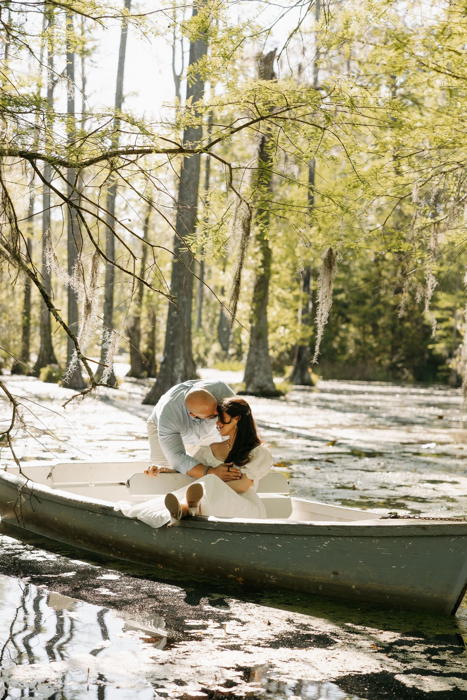 Couple sharing a romantic kiss in a white canoe surrounded by cypress trees in a serene swamp setting.