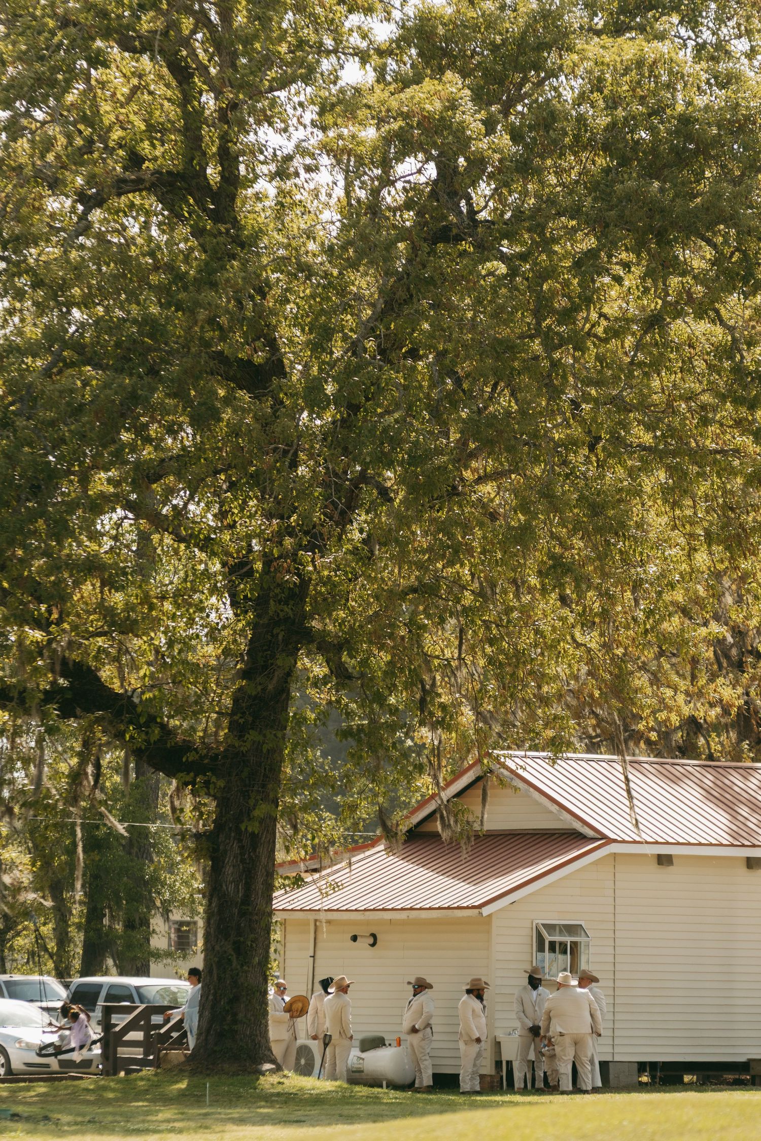 Wedding groomsmen in matching cream suits gathered outside white building beneath large oak tree.