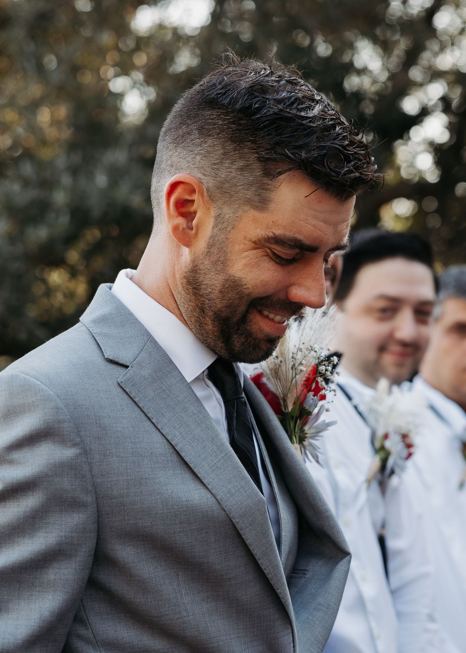 Smiling groom in a grey suit with a floral boutonniere standing outdoors at his wedding ceremony.