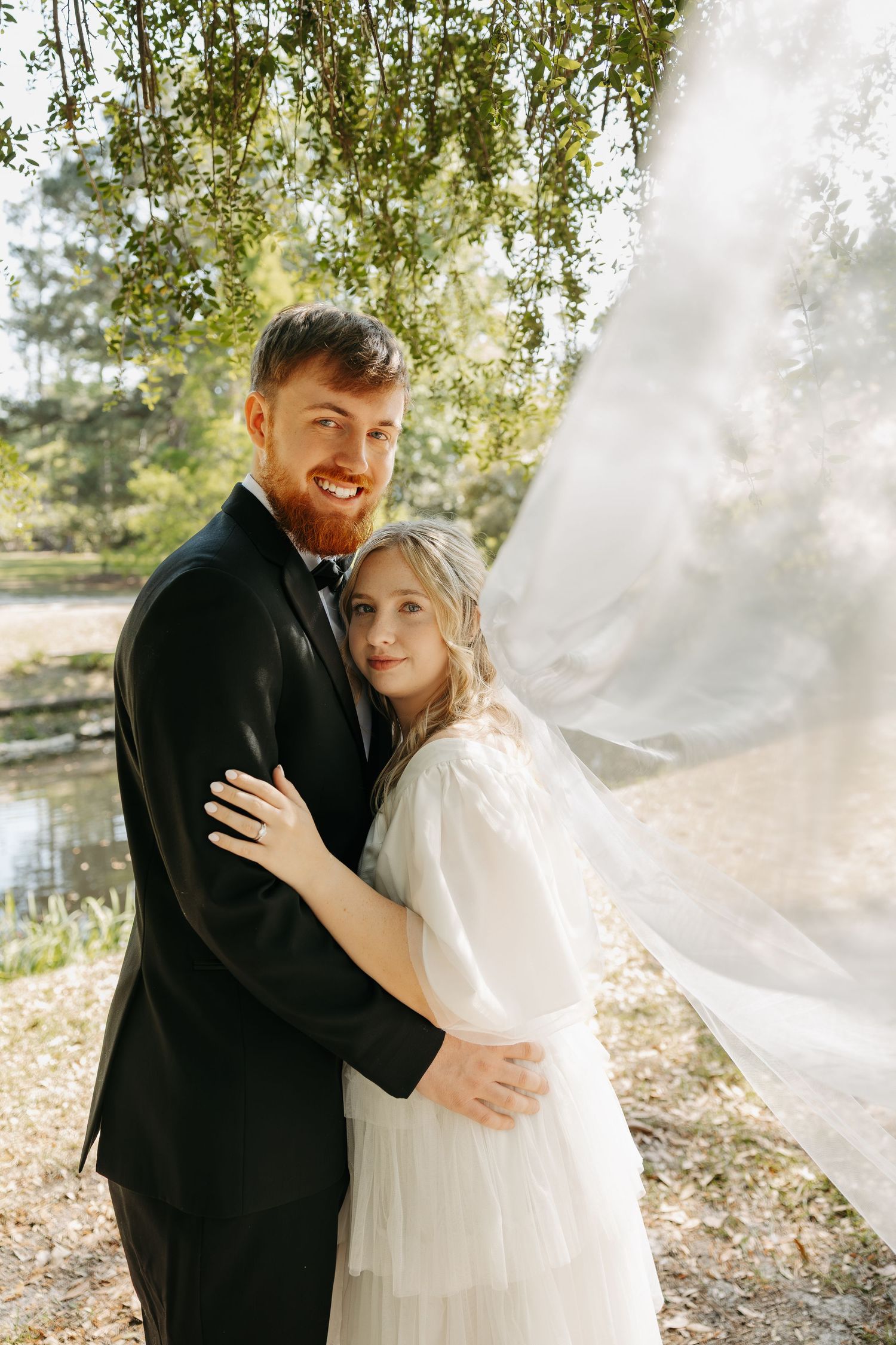Smiling groom in black tuxedo embraces bride in white dress with flowing veil in a sunlit outdoor garden setting.
