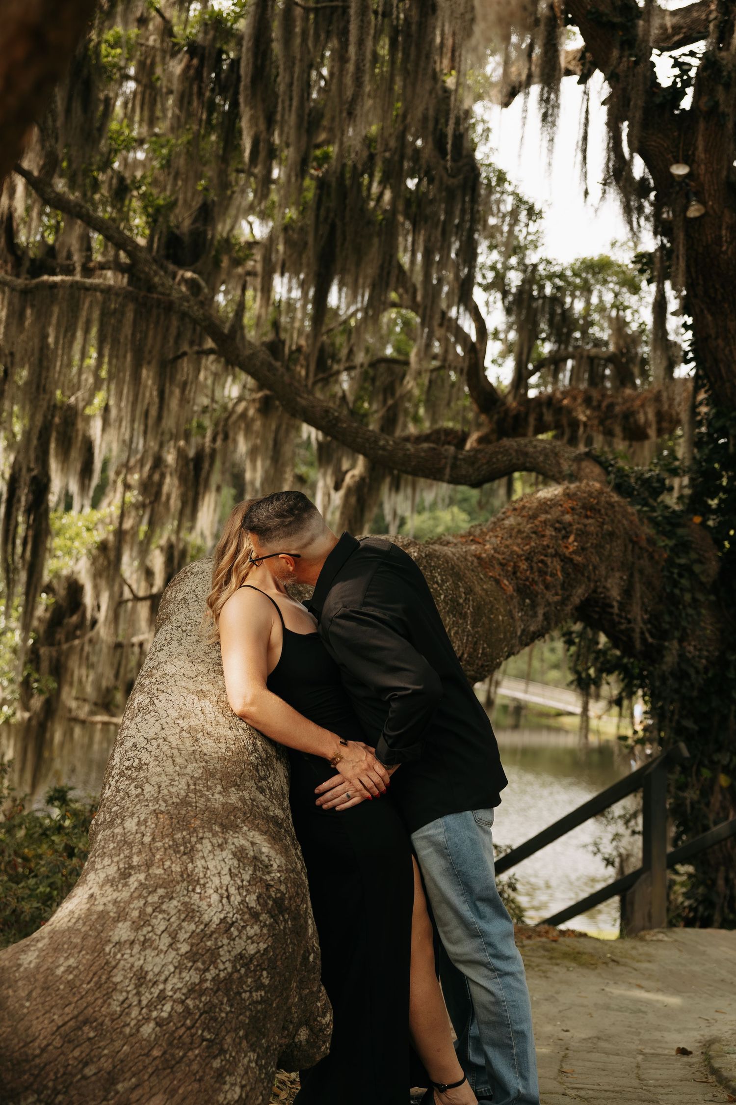 Couple embracing romantically against a moss-draped oak tree near a pond in a lush, atmospheric park setting.