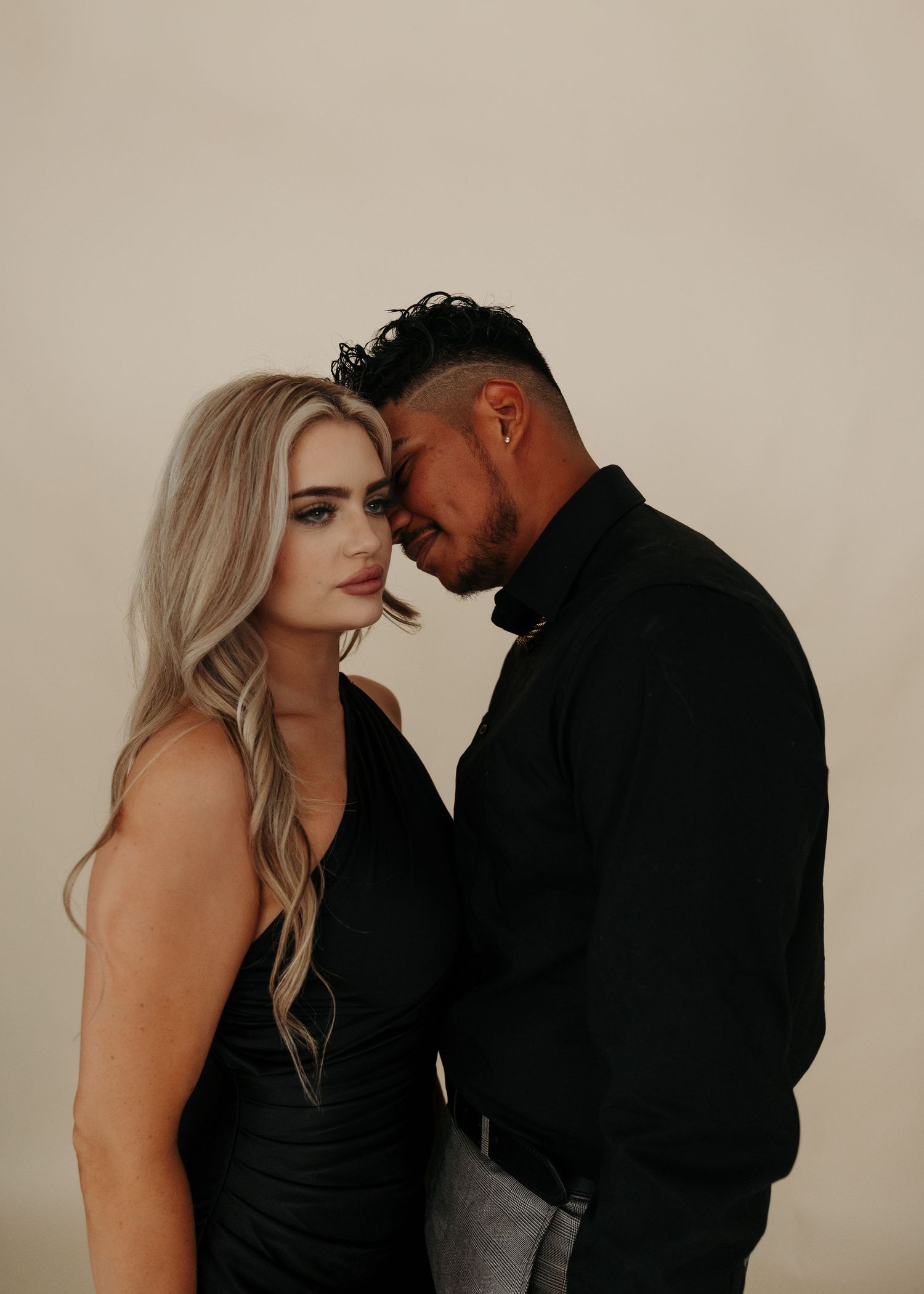 A couple in black outfits share an intimate moment, foreheads nearly touching, against a neutral background.