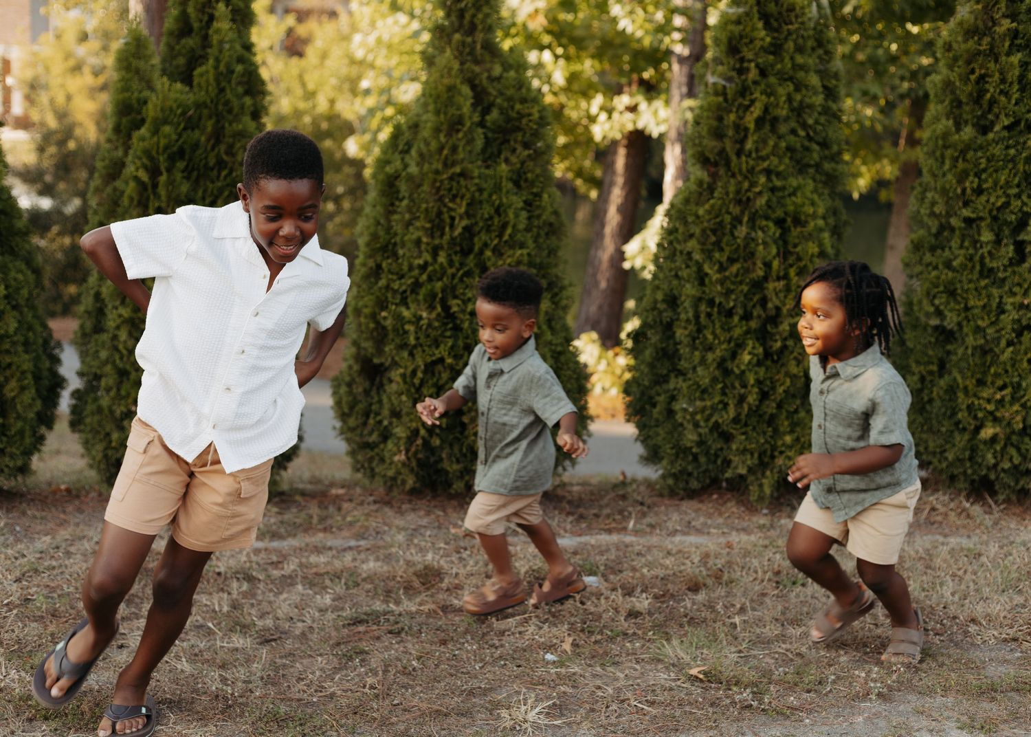Three young children playing and running outdoors in a park with trees in the background on a sunny day.