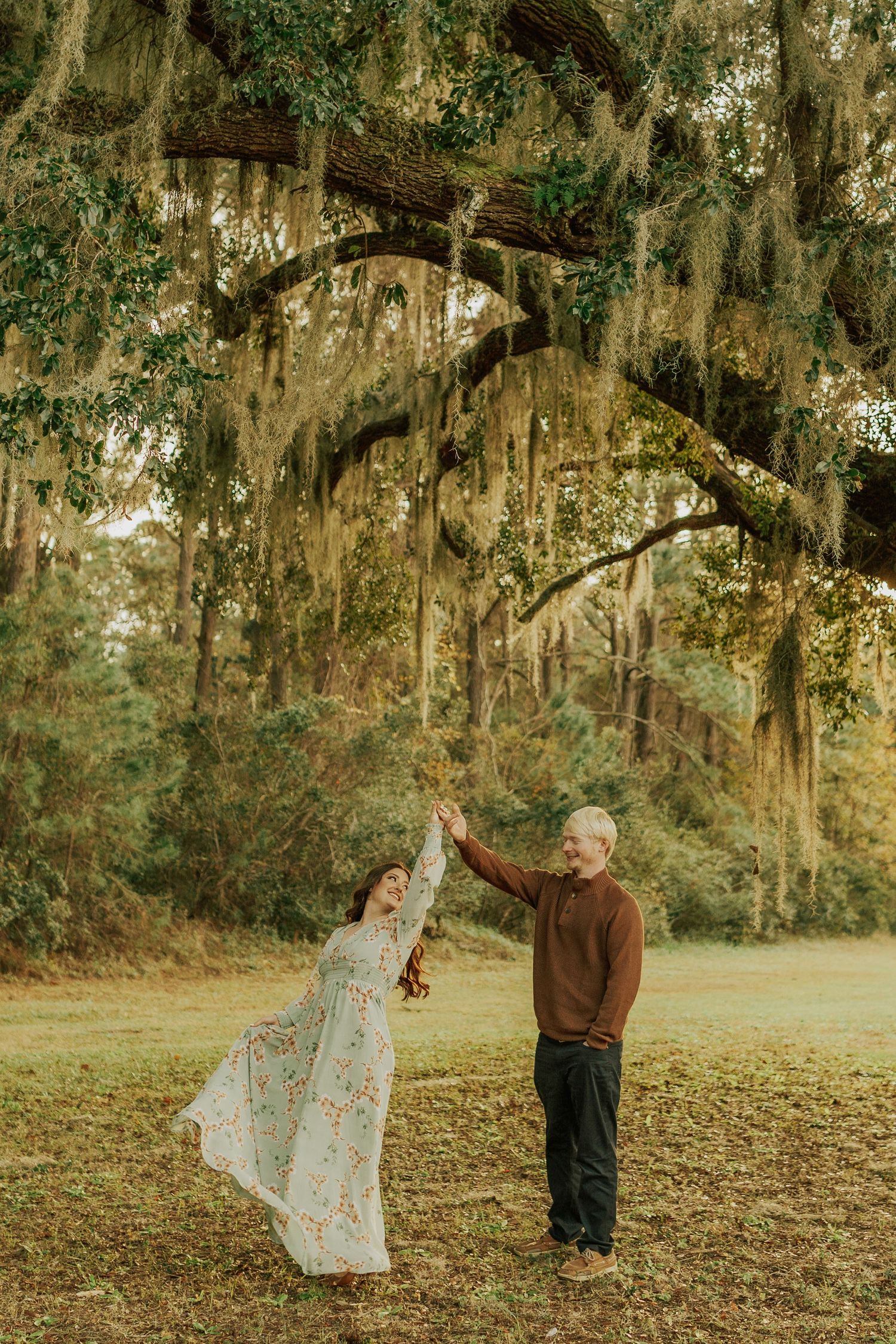 Couple dancing under Spanish moss-draped oak trees in a lush green park, woman in floral dress twirling.