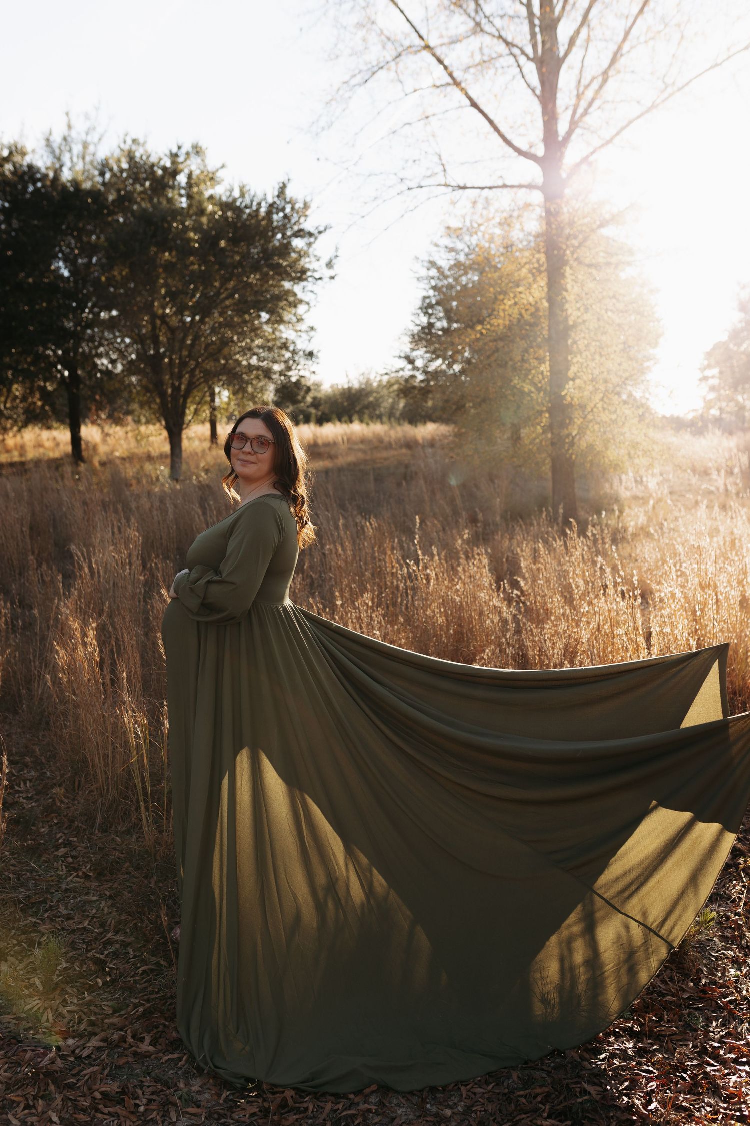 A figure in a flowing olive green dress stands in a sunlit autumn field with tall grass and trees at golden hour.