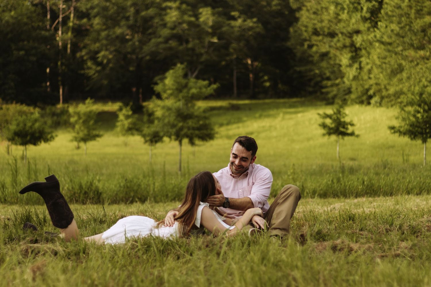 A couple embraces in a romantic pose on a grassy hillside surrounded by lush green trees on a sunny day.