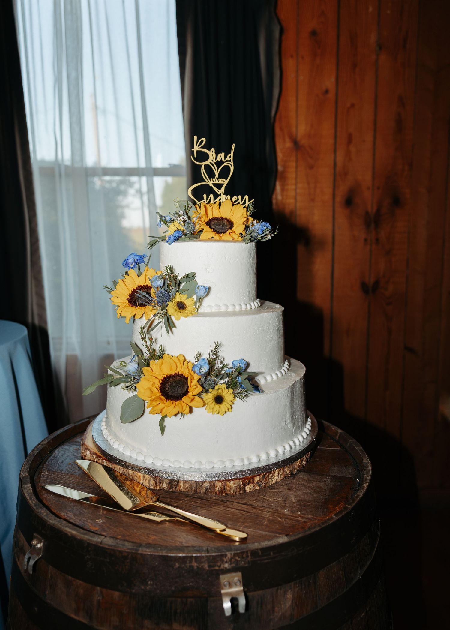 Three-tier white wedding cake with sunflowers and blue flowers, topped with custom name topper, displayed on a rustic wooden barrel.