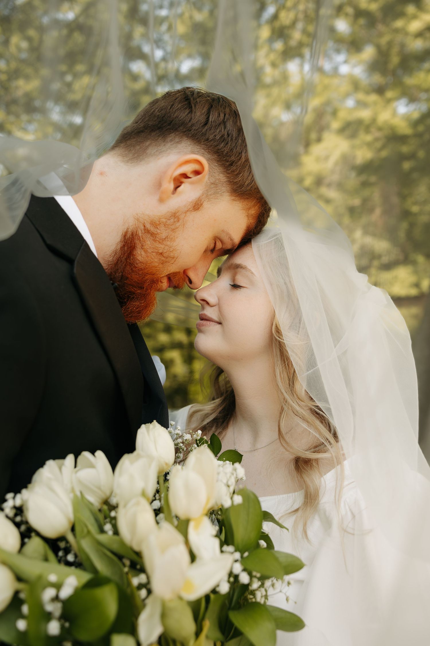 Bride and groom touching foreheads under veil, holding white tulip bouquet, romantic outdoor wedding portrait.
