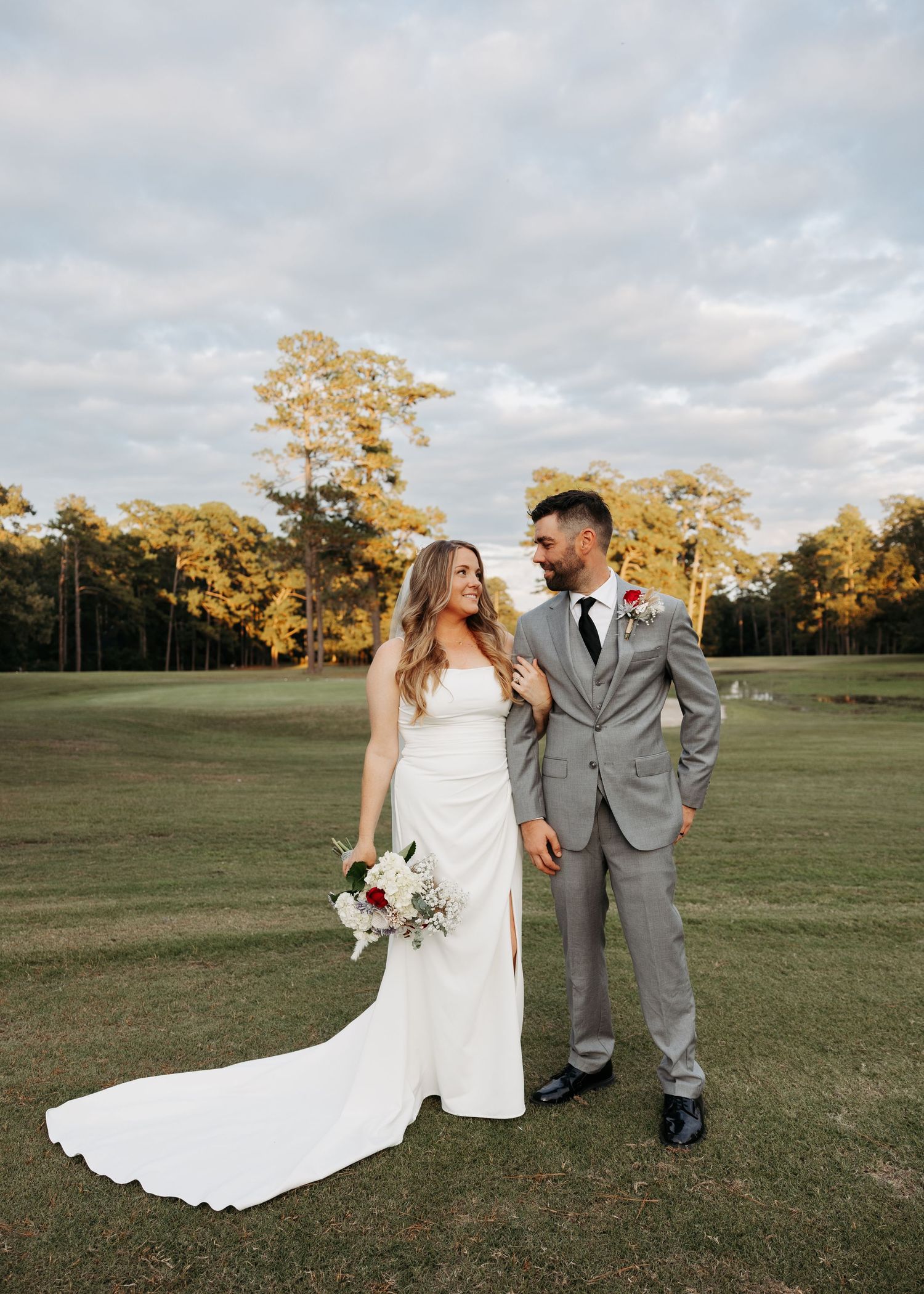Bride in white strapless gown and groom in gray suit posing together on a golf course at golden hour.