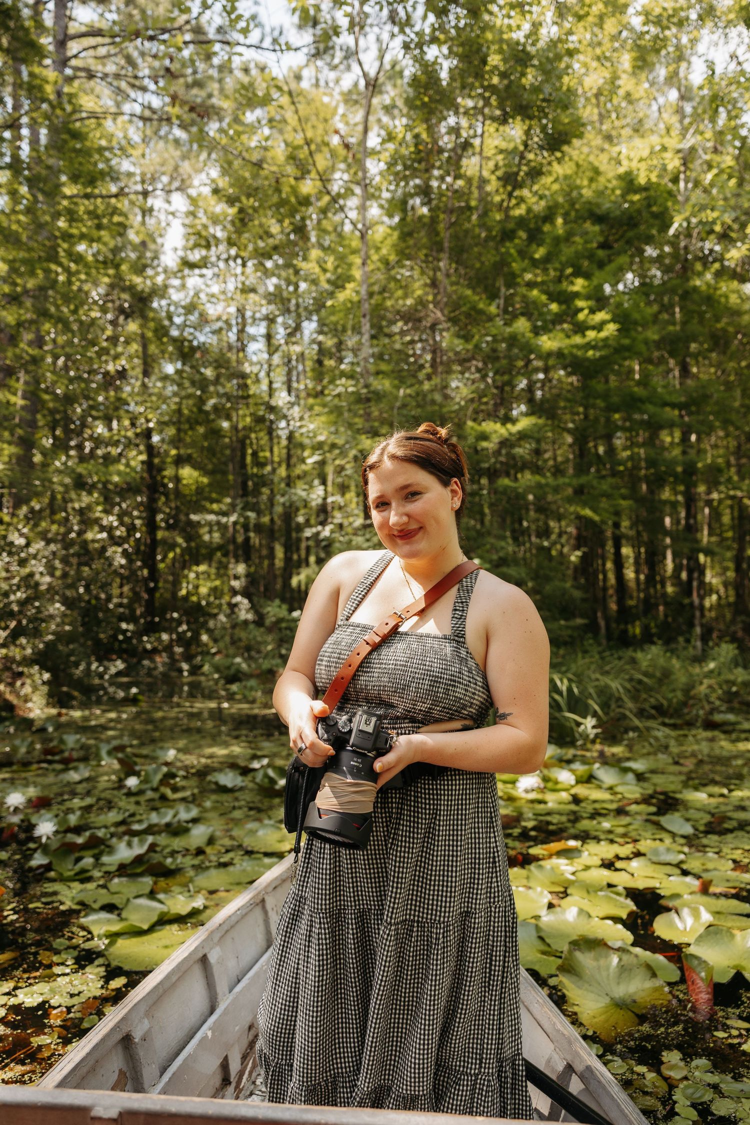 Woman in checkered dress holding camera stands in canoe surrounded by lily pads and lush green forest.