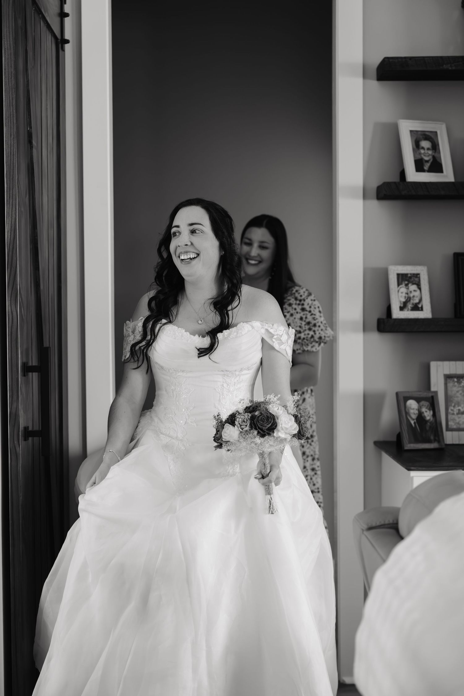 Black and white photo of a laughing bride in a wedding gown holding a bouquet, with a smiling woman standing behind her.