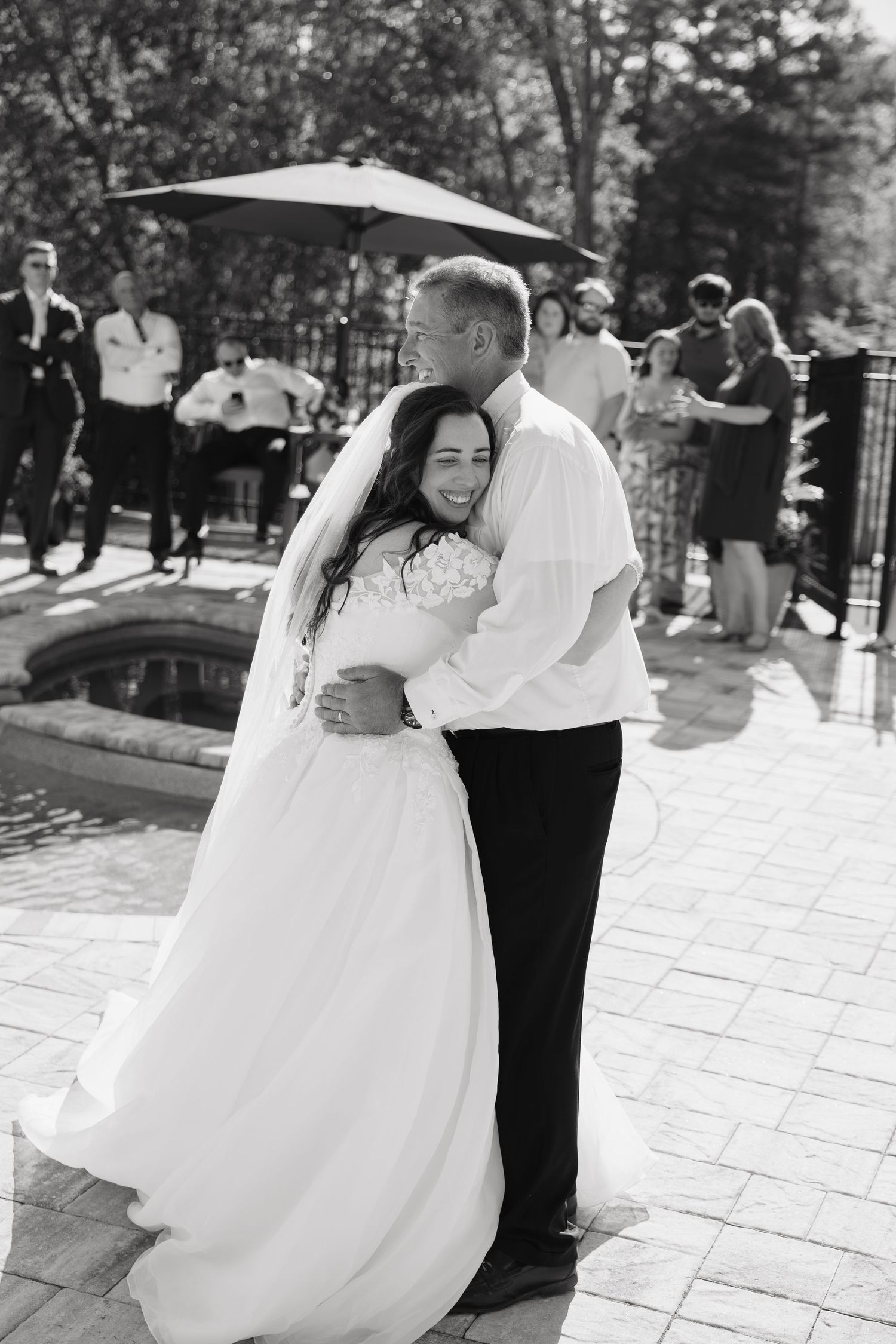 Black and white photo of bride and groom sharing first dance outdoors at wedding reception with guests in background.