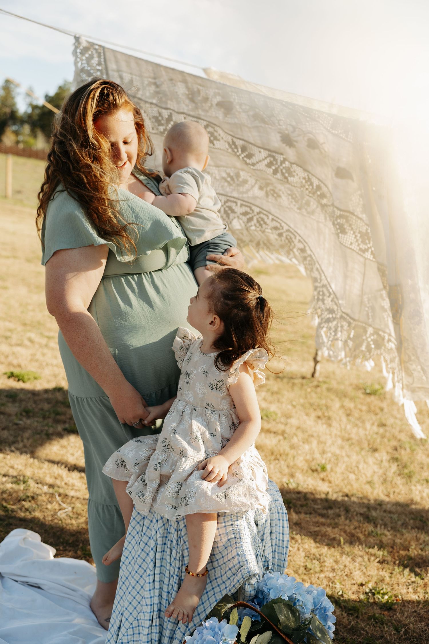 Pregnant woman in green dress holding baby while toddler girl in floral dress hugs her belly outdoors.