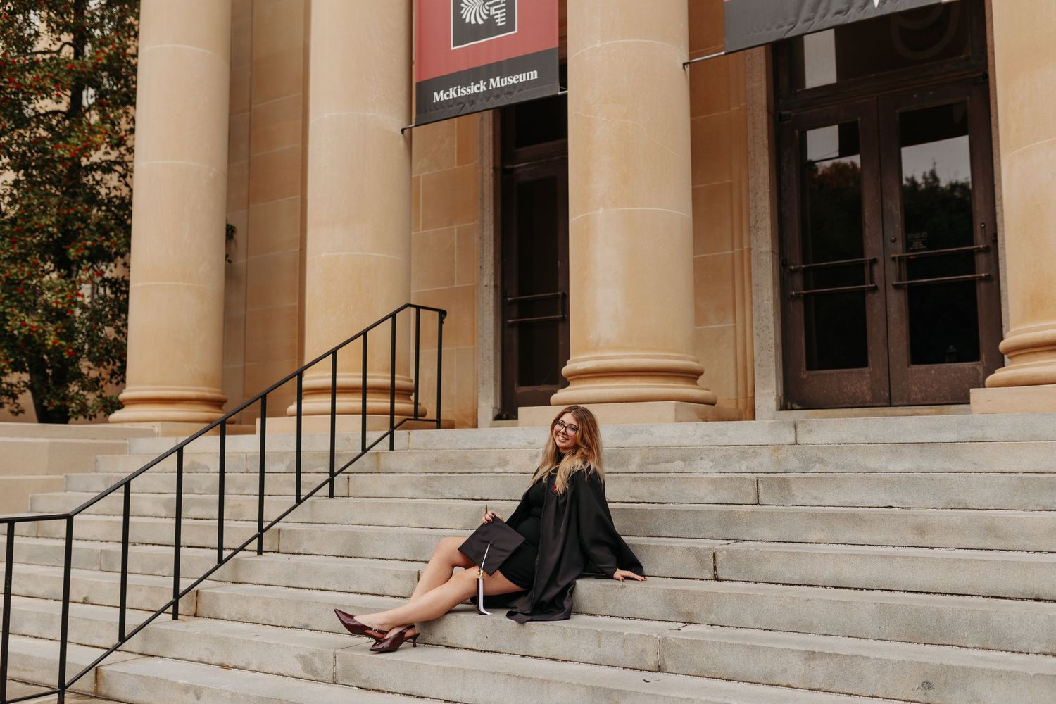 A smiling woman in a black graduation gown sits on steps outside a classical building with tall columns.