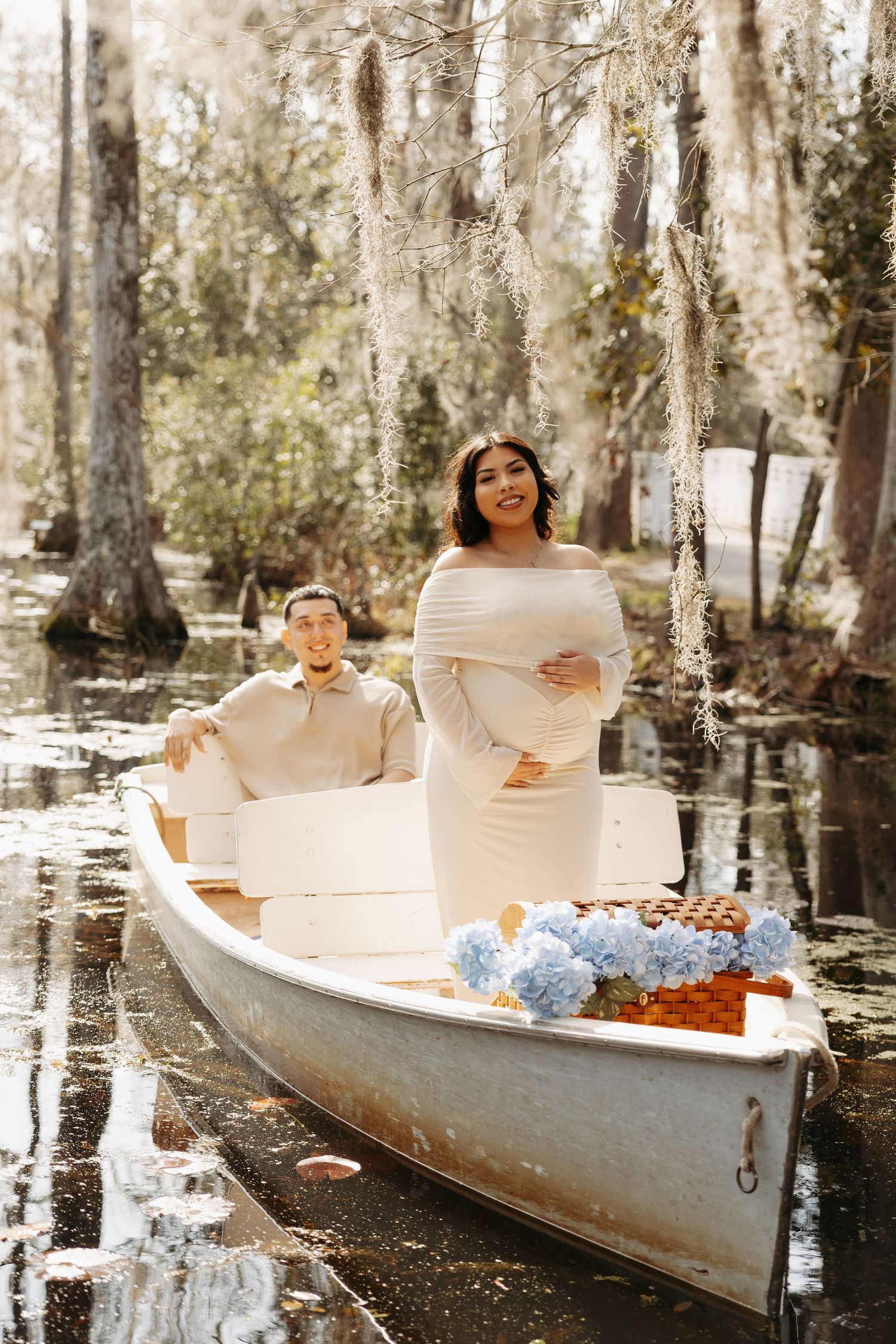 Pregnant woman in white dress stands in a boat with her partner amid a serene cypress swamp, blue hydrangeas nearby.