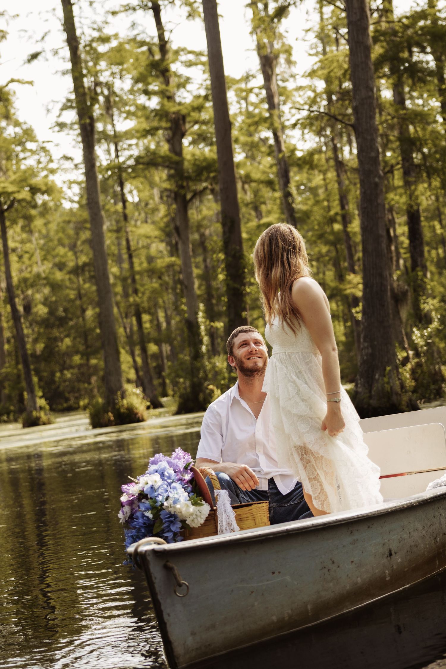 Couple in a rowboat on a serene lake surrounded by tall trees, woman in white dress standing as man smiles up at her.