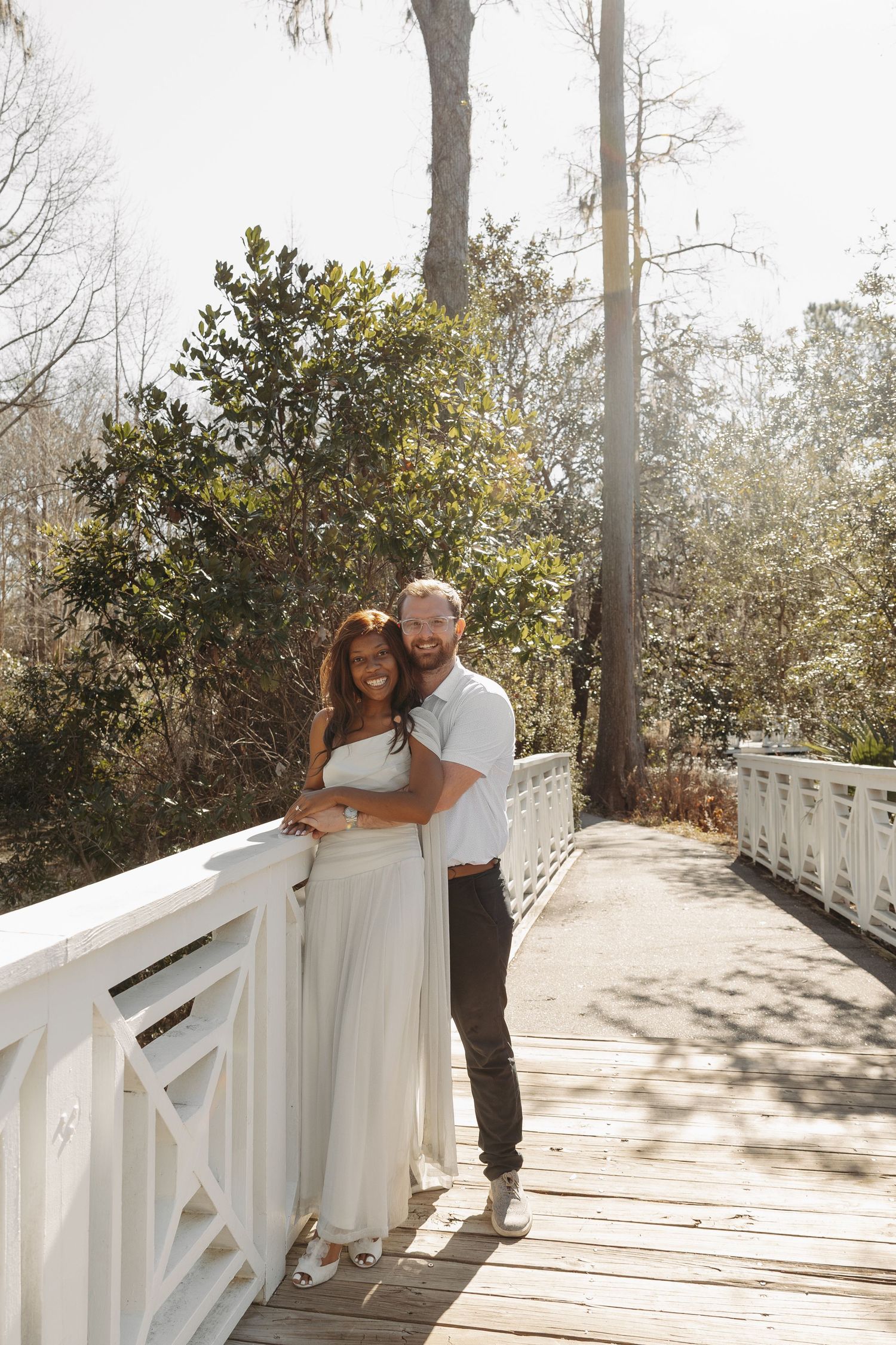 Couple embracing on a white wooden bridge surrounded by lush greenery on a sunny day.