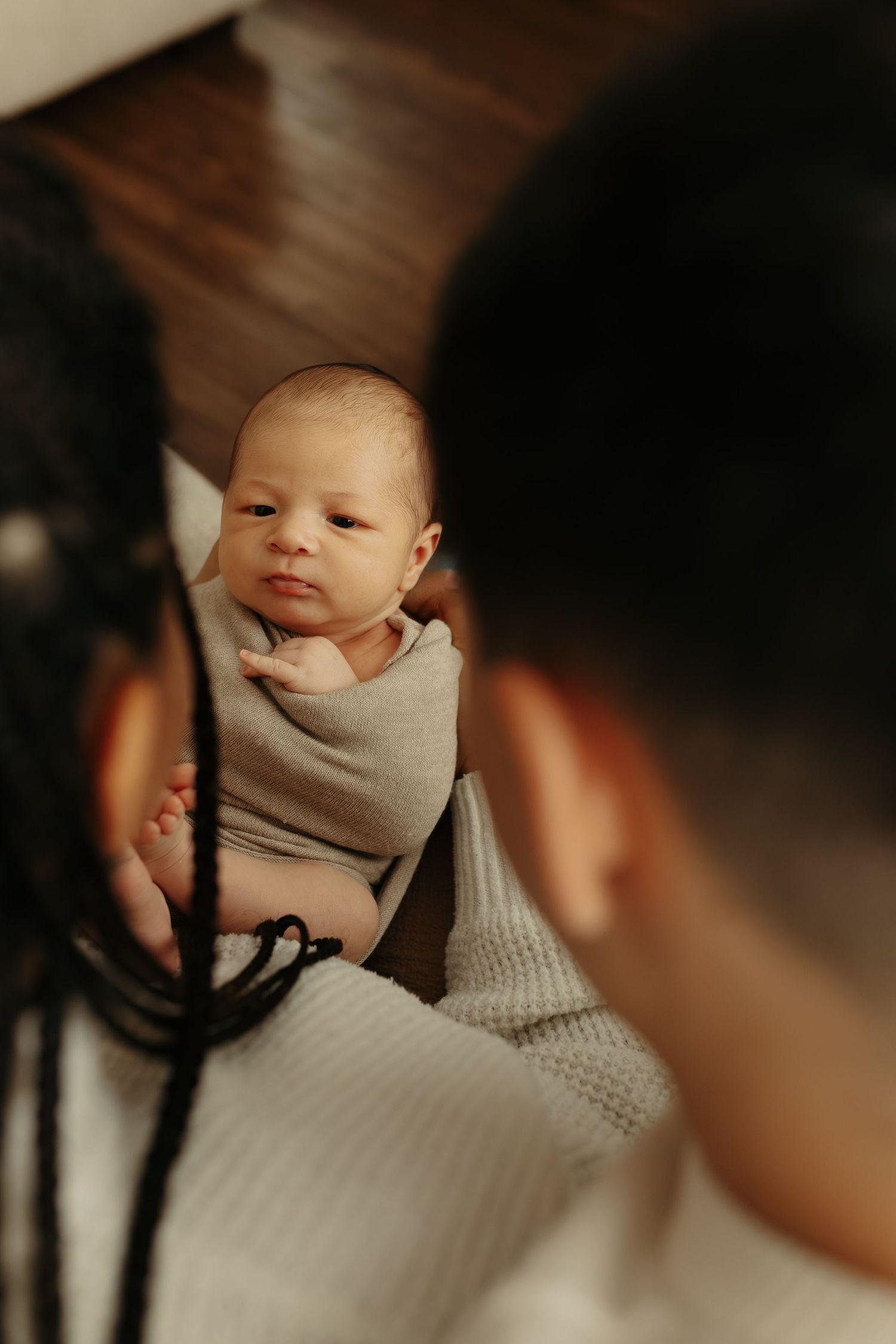 A newborn baby wrapped in a gray swaddle blanket looks off to the side during an intimate indoor photo session.