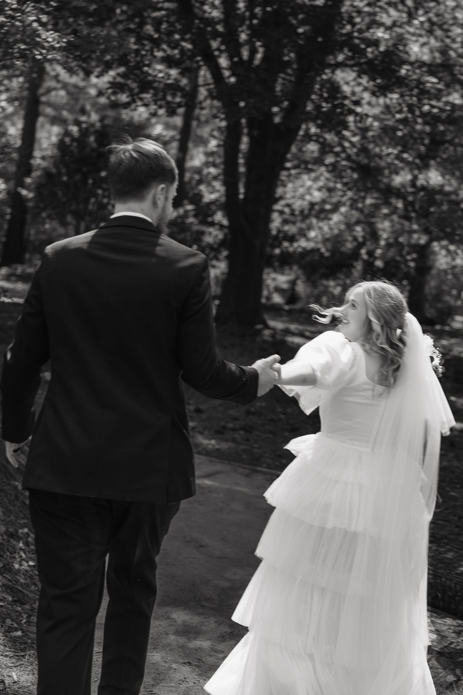 Black and white photo of a bride and groom walking hand in hand through a lush green forest path.