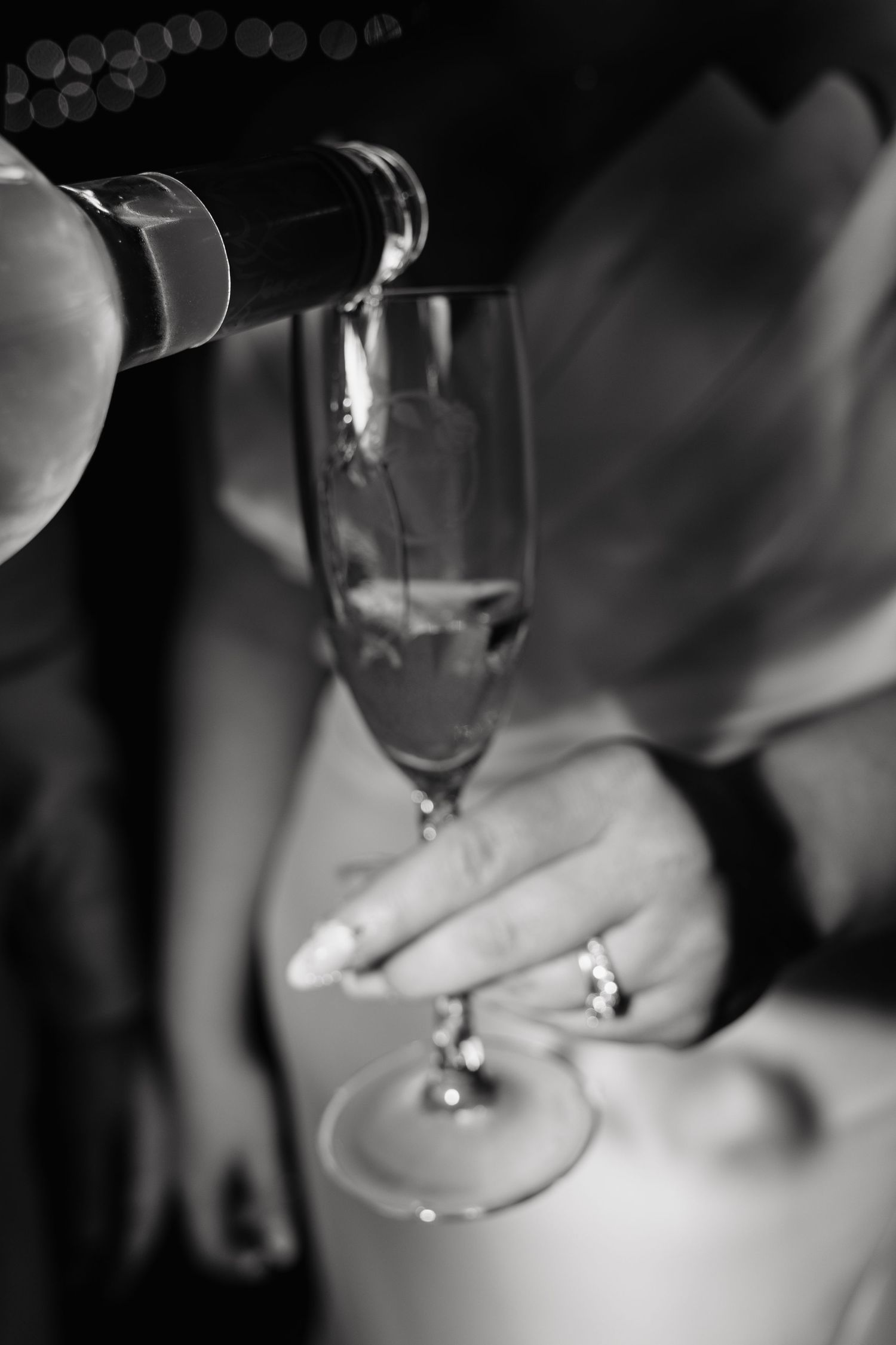 Champagne being poured into flute held by bride wearing engagement ring, black and white photo.