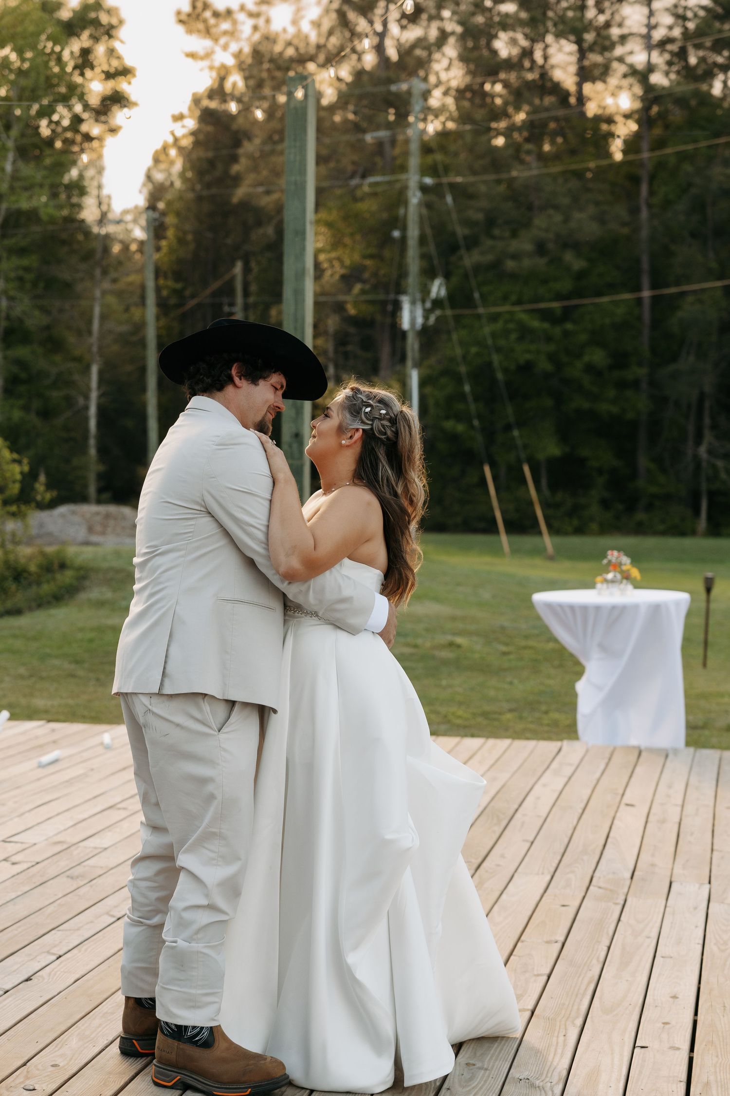 Bride and groom share a romantic first dance on a wooden deck surrounded by lush greenery at sunset.