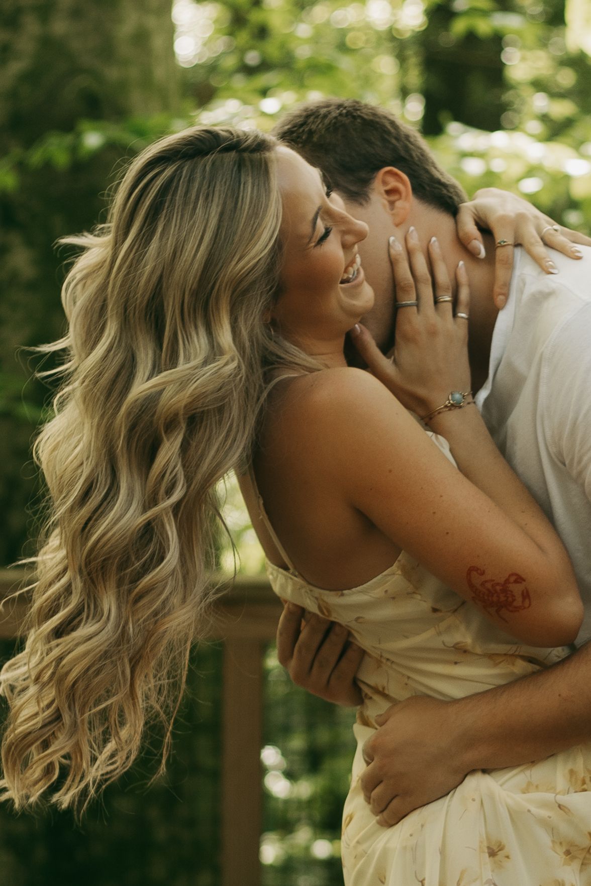 A smiling woman with long blonde wavy hair embraces a man outdoors, wearing a floral dress with a heart tattoo on her arm.