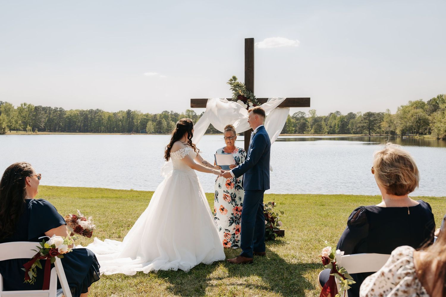 Outdoor wedding ceremony by a lake with bride and groom holding hands before a wooden cross altar.