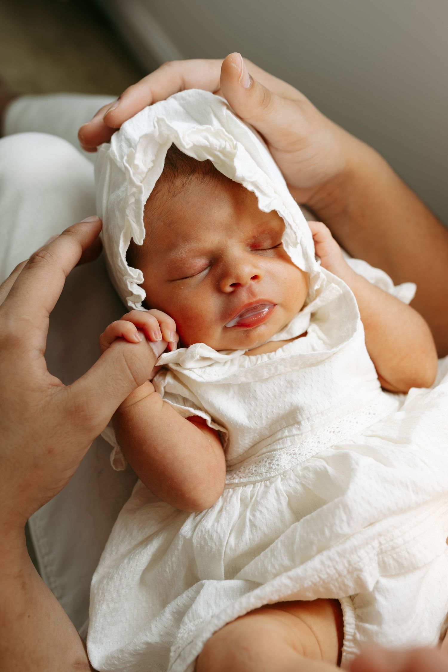 Newborn baby in white bonnet and dress sleeping peacefully while being held by adult hands.