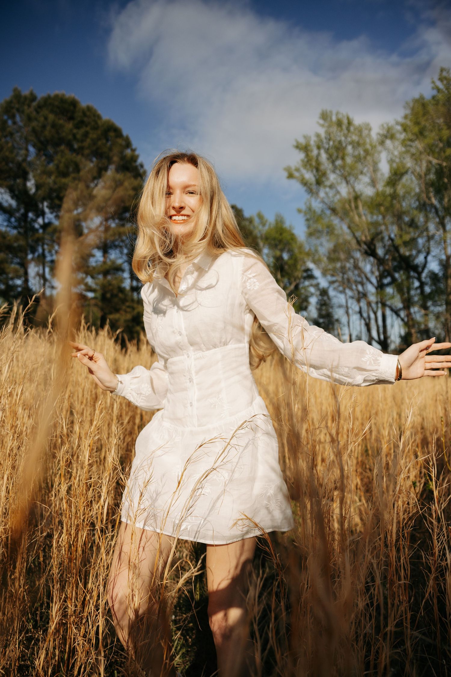 Smiling blonde woman in white dress standing in golden wheat field with trees and blue sky background.