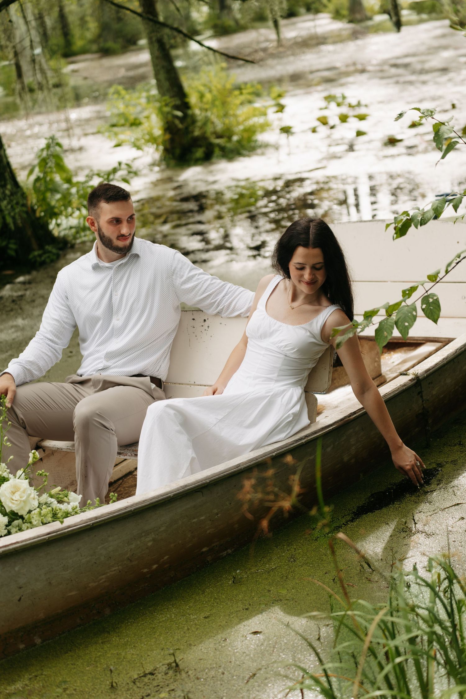 Bride and groom in white attire sitting in a decorated boat on a serene green swamp.