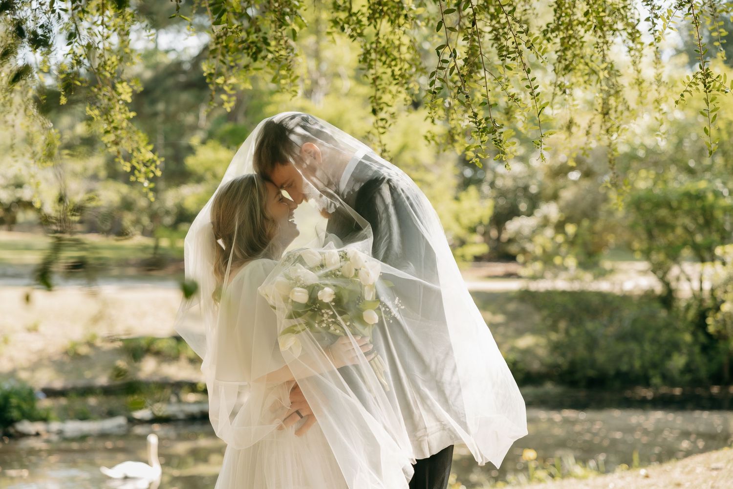 Bride and groom share a romantic kiss under a weeping willow tree, with bridal veil flowing and white bouquet in hand.
