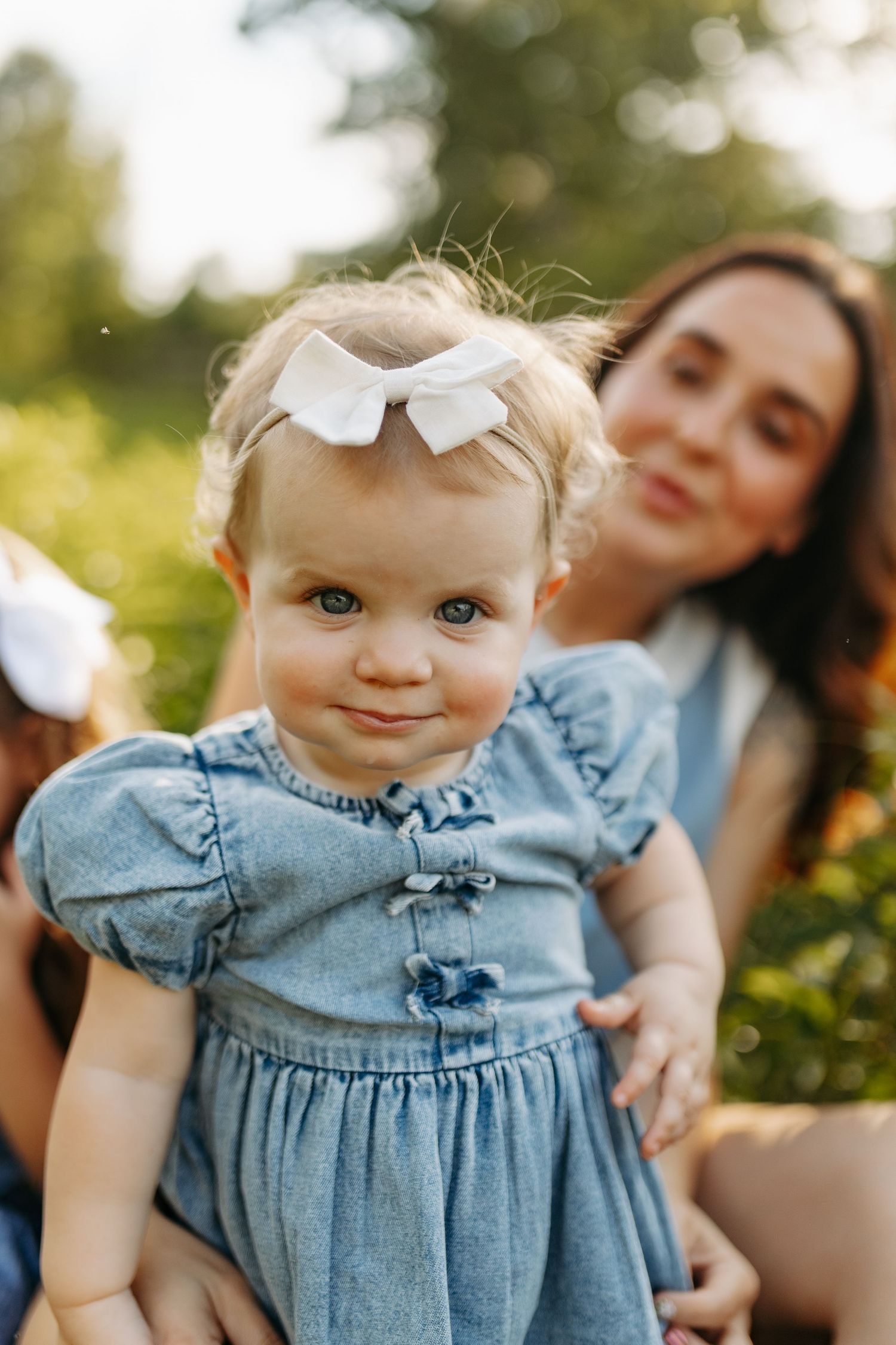 Adorable baby girl in blue denim dress with white bow smiles outdoors, held by her brunette mother in sunny park.