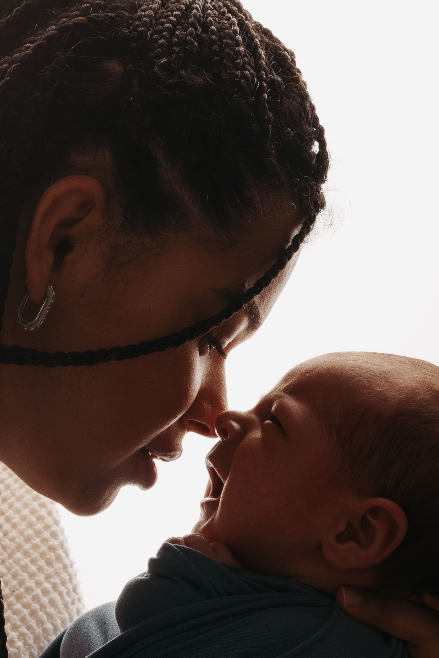 A tender moment between a young Black mother with braids and her newborn baby touching noses in a warm backlit portrait.