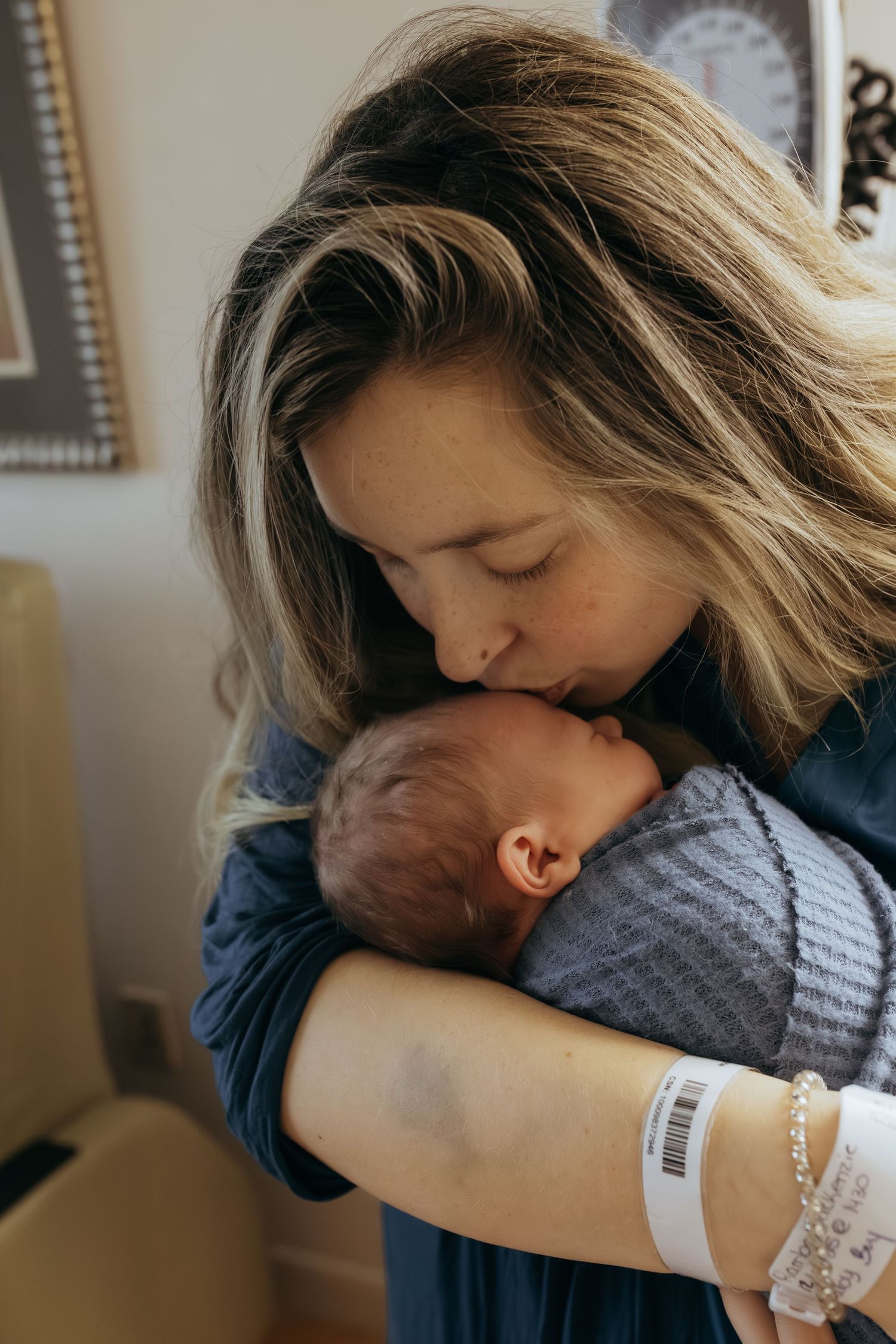 New mother tenderly kissing her newborn baby while holding it in her arms, wearing a hospital ID bracelet.