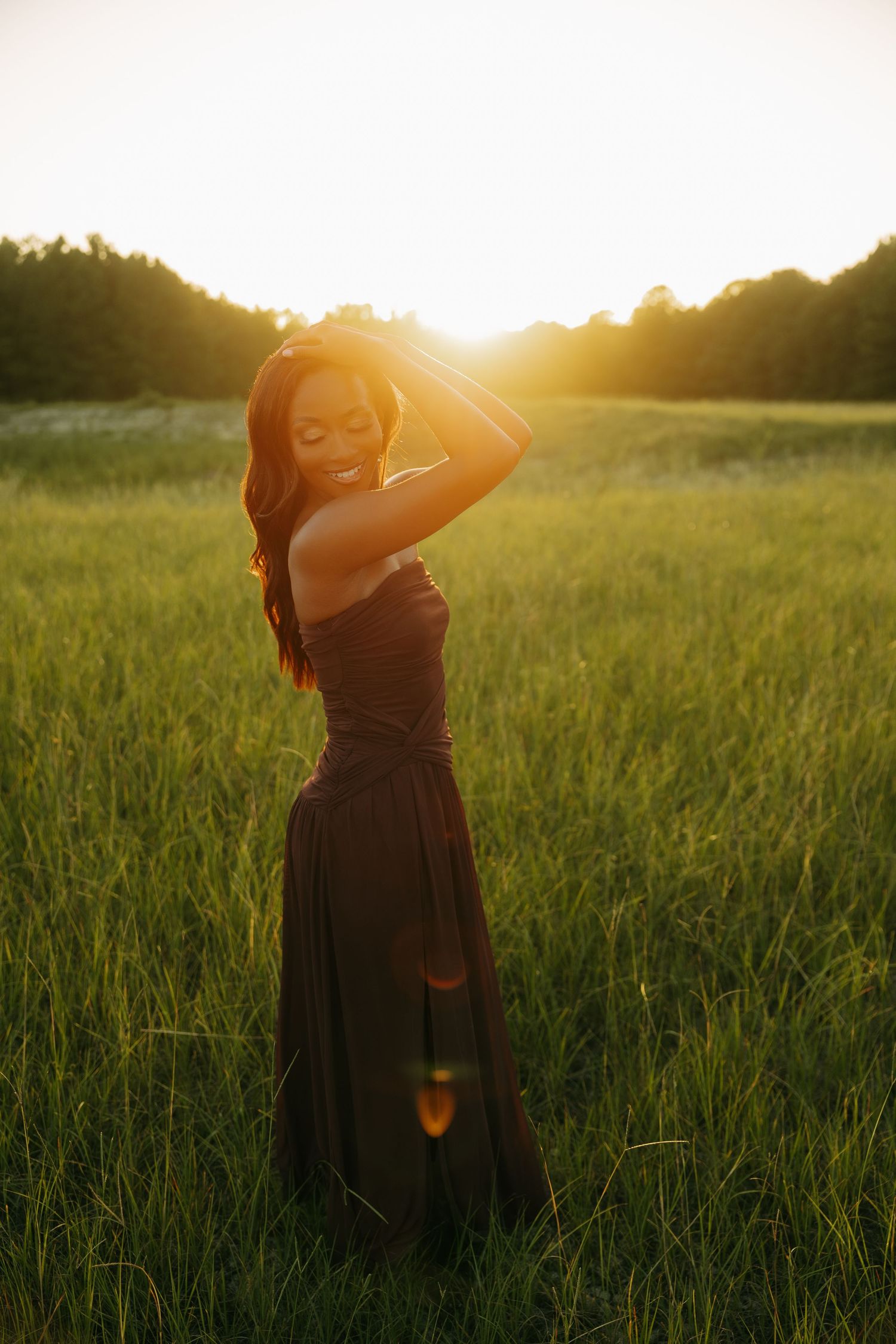 Woman in dark gown standing in sunlit green field at golden hour with warm backlight and lens flare.