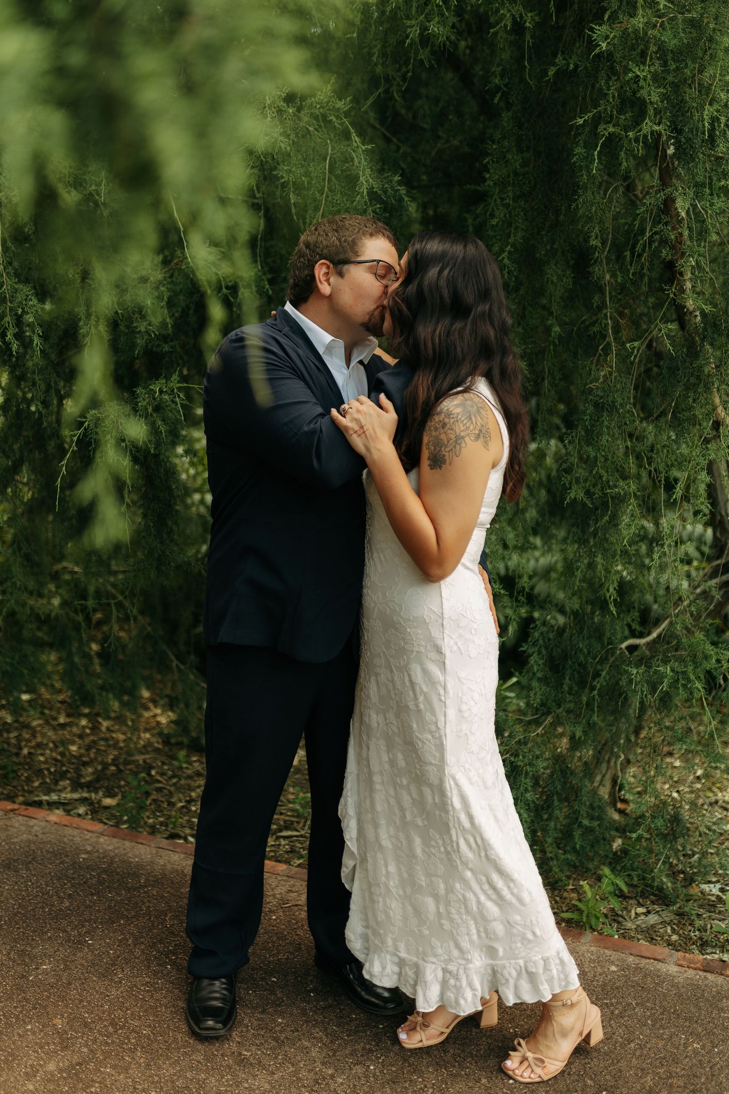 A couple shares a romantic kiss outdoors, wearing wedding attire, surrounded by lush green trees.