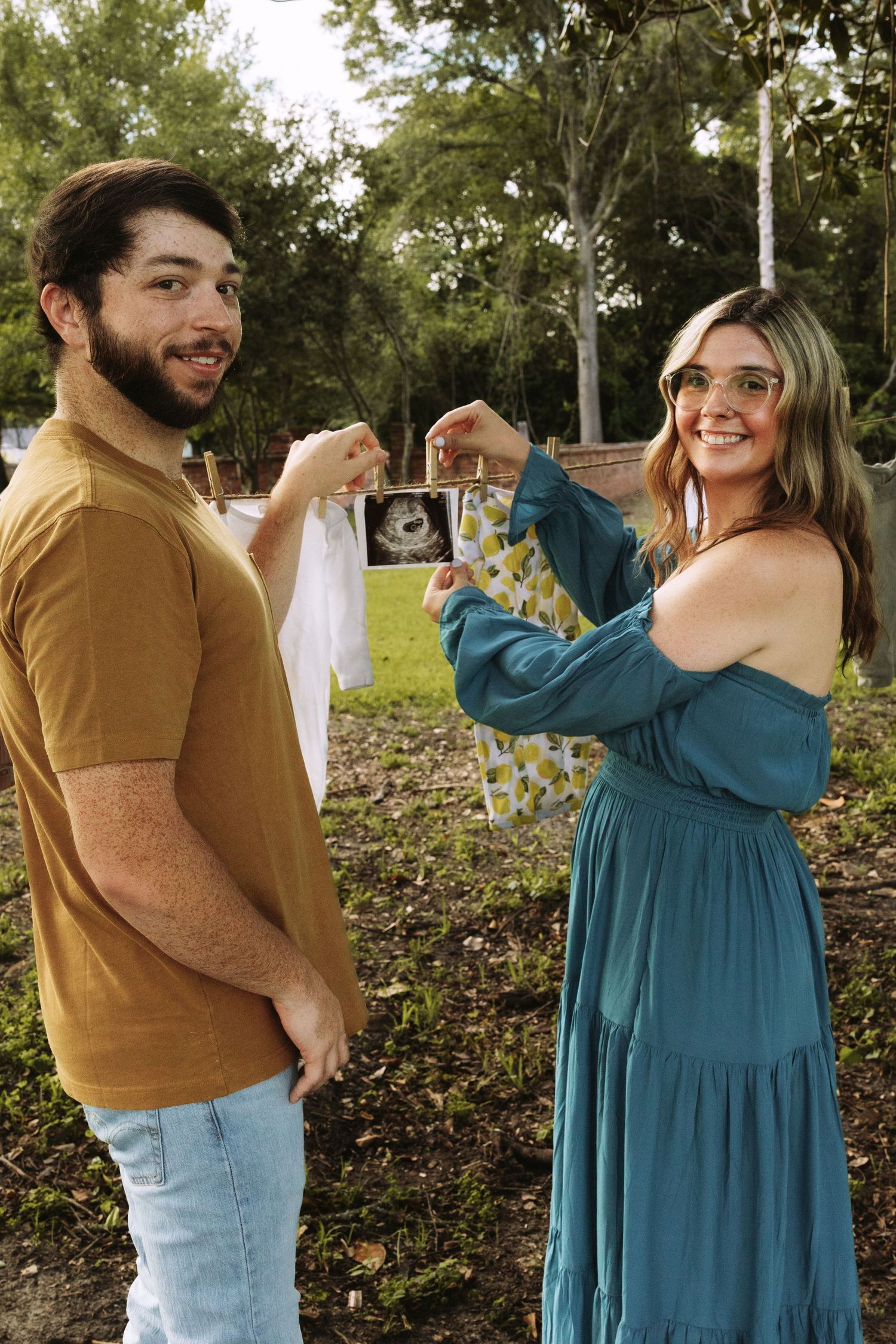 A couple in casual attire holds up an ultrasound photo together while standing outdoors in a natural setting.