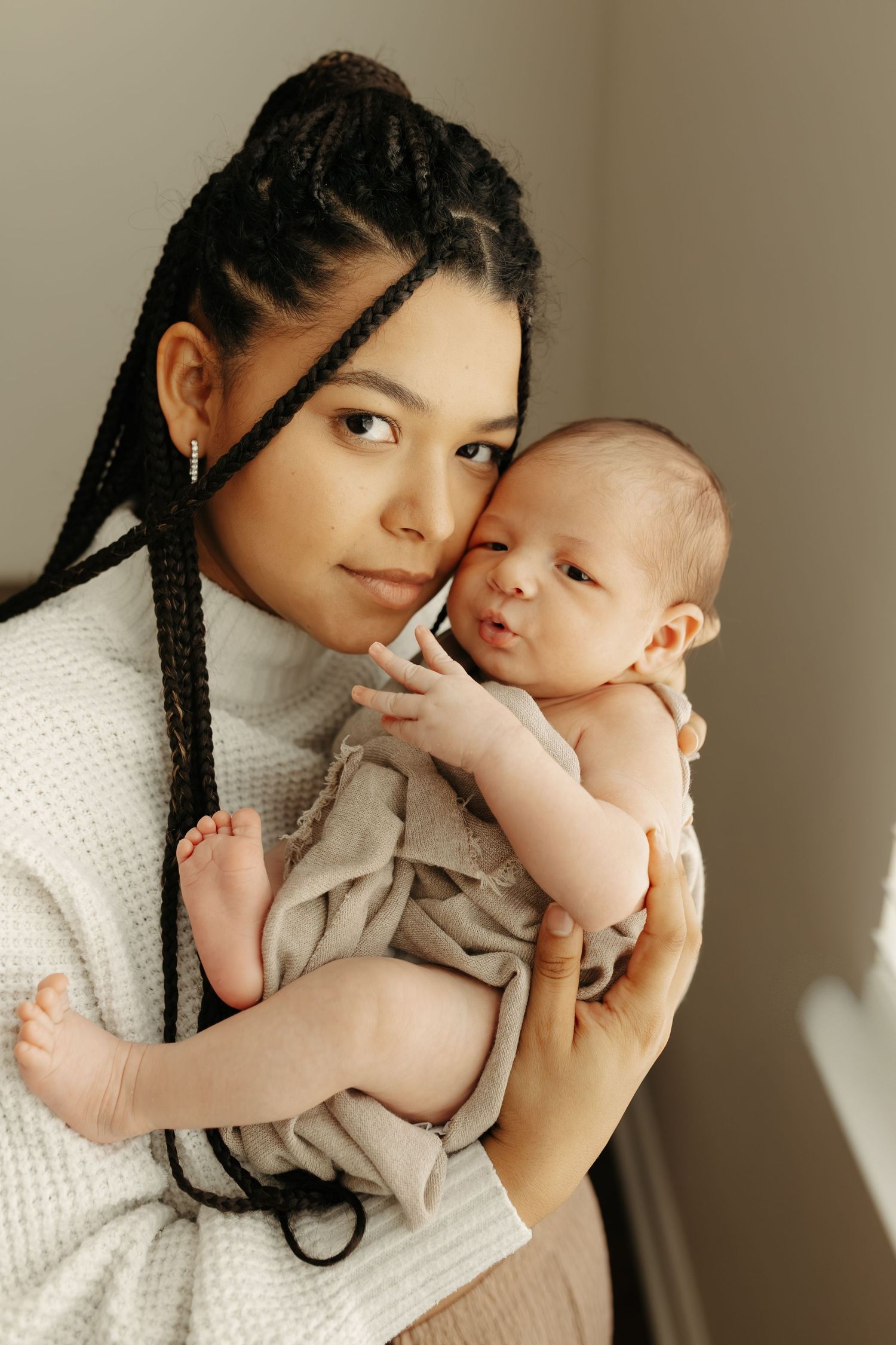 A young mother with box braids tenderly holds her newborn baby close to her cheek, both dressed in neutral tones.