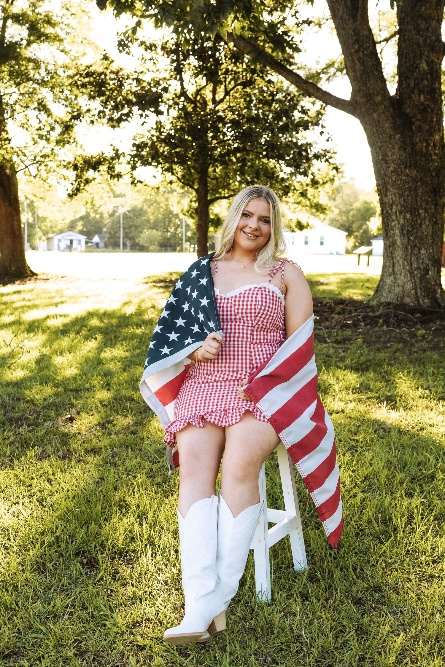 Woman in red gingham dress and boots sits with American flag on grass in golden sunlight.
