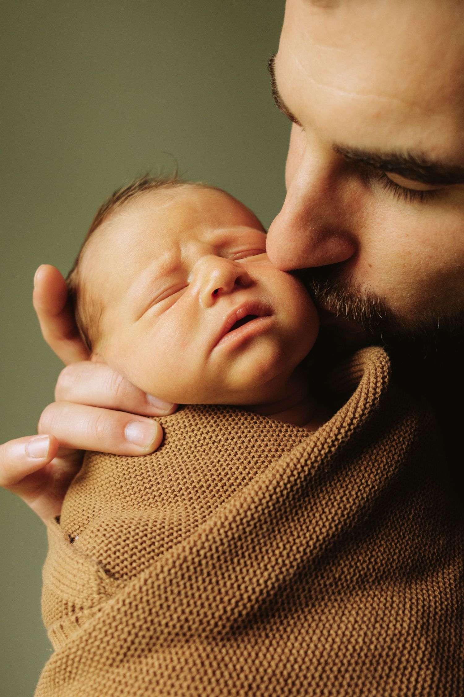 A tender moment captured as a bearded person gently embraces a sleeping newborn wrapped in a brown knit blanket.